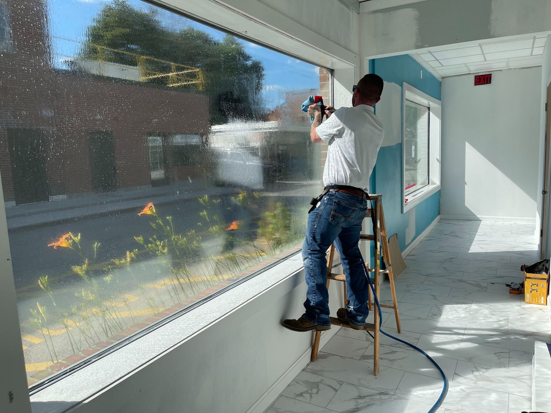 A person on a ladder installs window film in a building with large windows and blue walls.