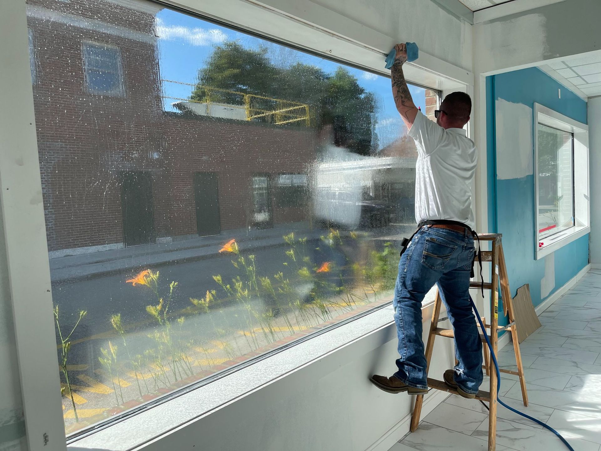 Man cleaning a large window from a ladder, inside a building under construction.