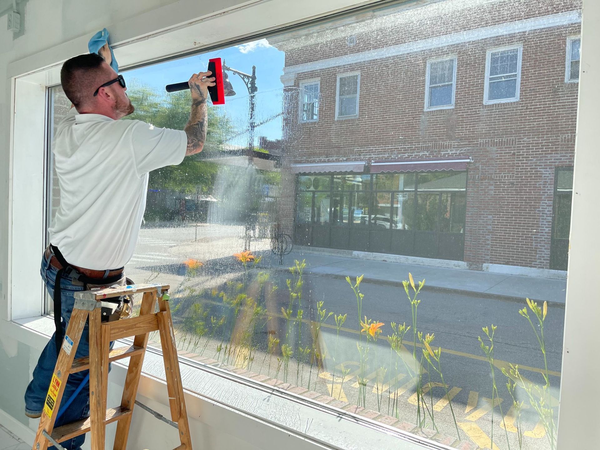 Man on a ladder applying tint to a large window with squeegee; downtown setting visible outside.