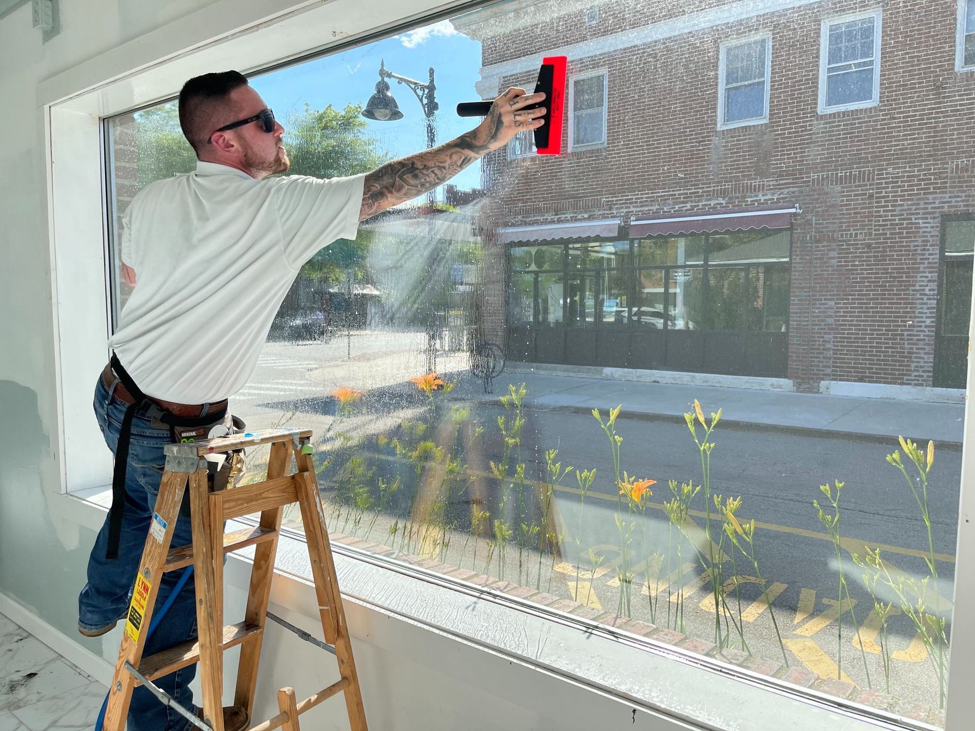 Person applies window film to a large window with a squeegee. A street and brick building are visible outside.