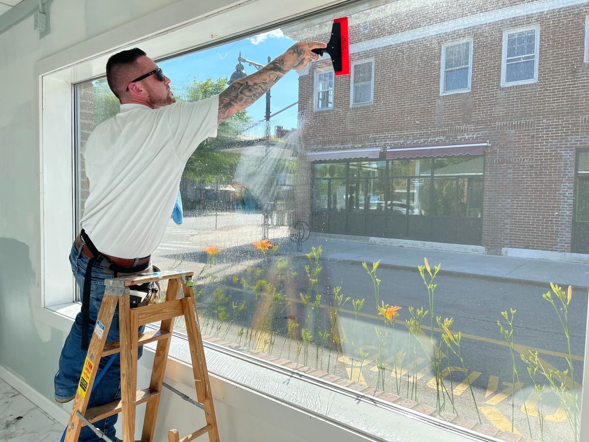 Man on a ladder washing a large window with a squeegee; downtown setting with a brick building in the background.