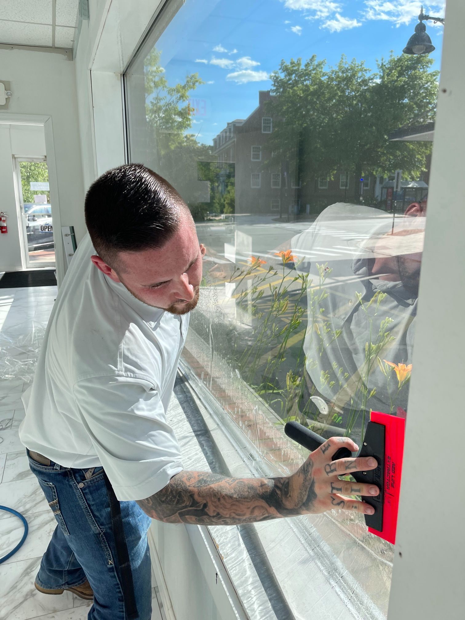 Man in white shirt cleaning a large window with a squeegee; outdoors background, sunny.