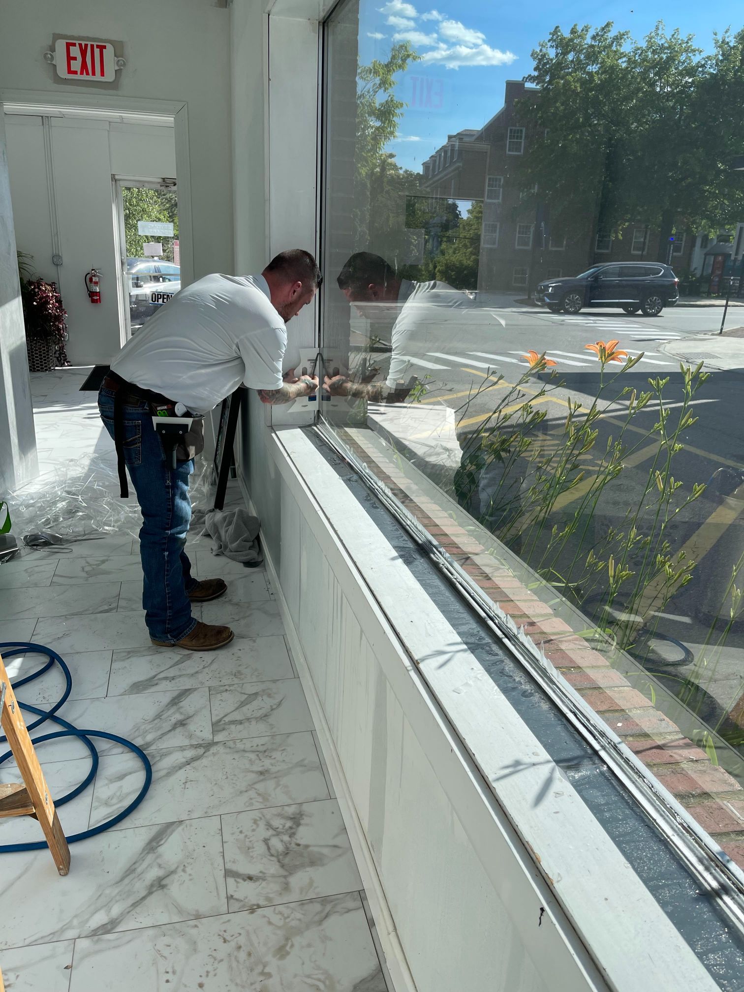 Man working on a storefront window, using tools. The window overlooks a street with parked cars and a building.