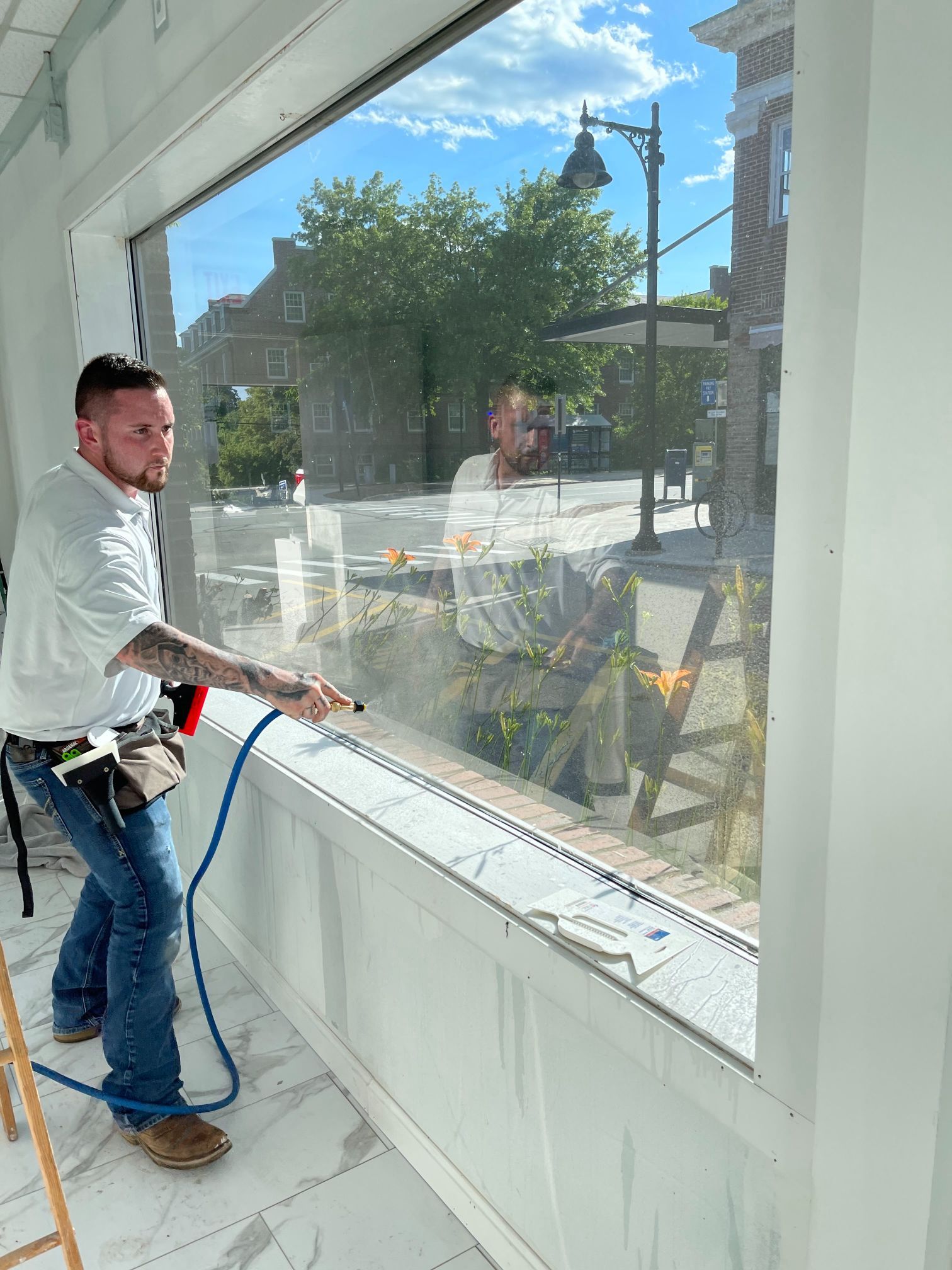 Man spraying window with blue hose, street and buildings reflected in glass.
