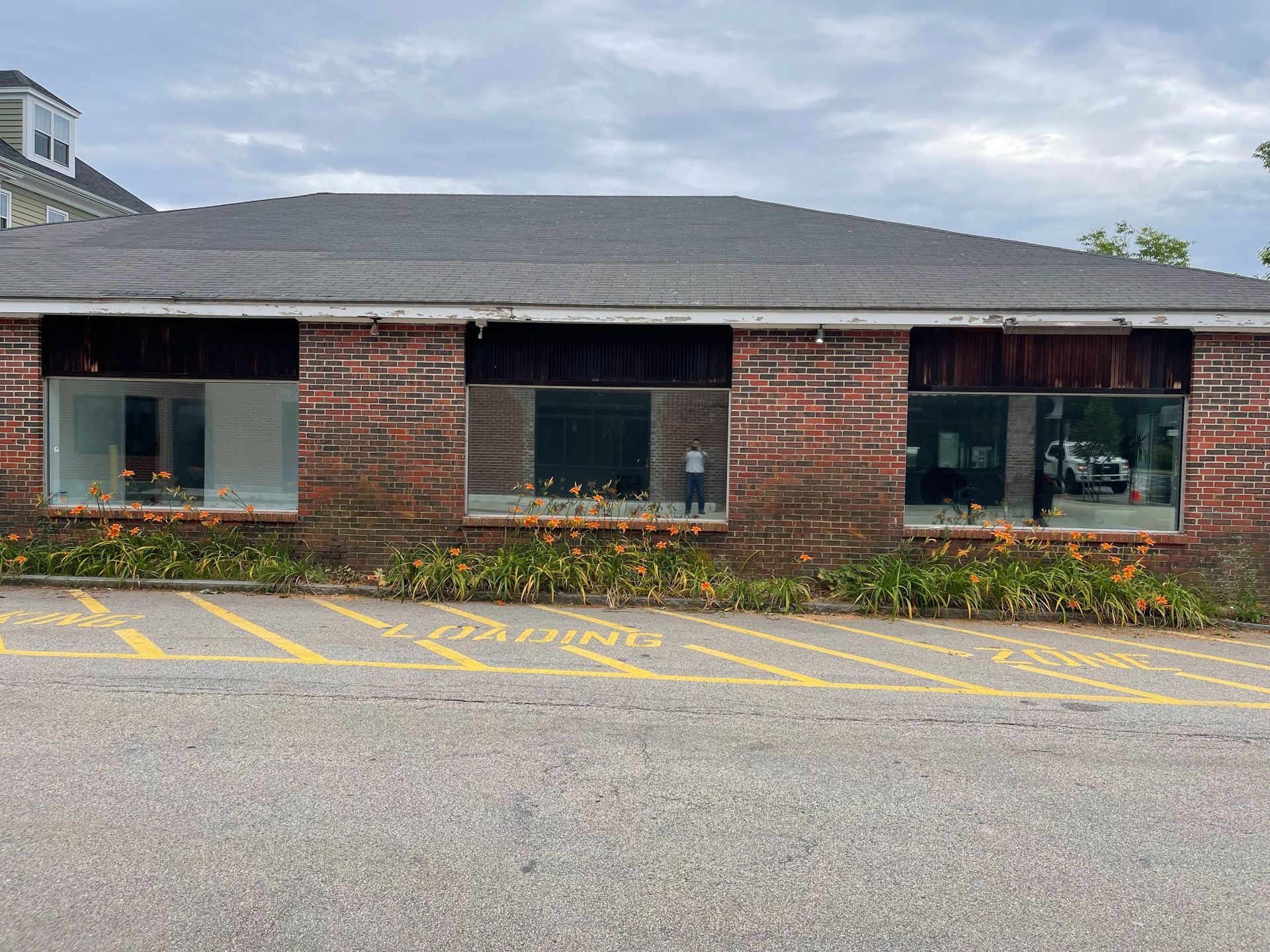 Brick building with three large windows; parking spaces in front, yellow flowers. Cloudy sky.