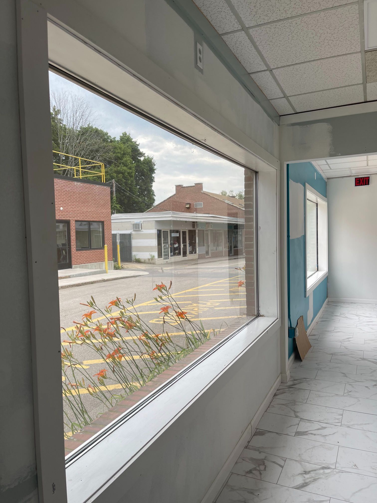 Interior view through a large window. Street scene visible, with a brick building and plants. Light blue walls and 