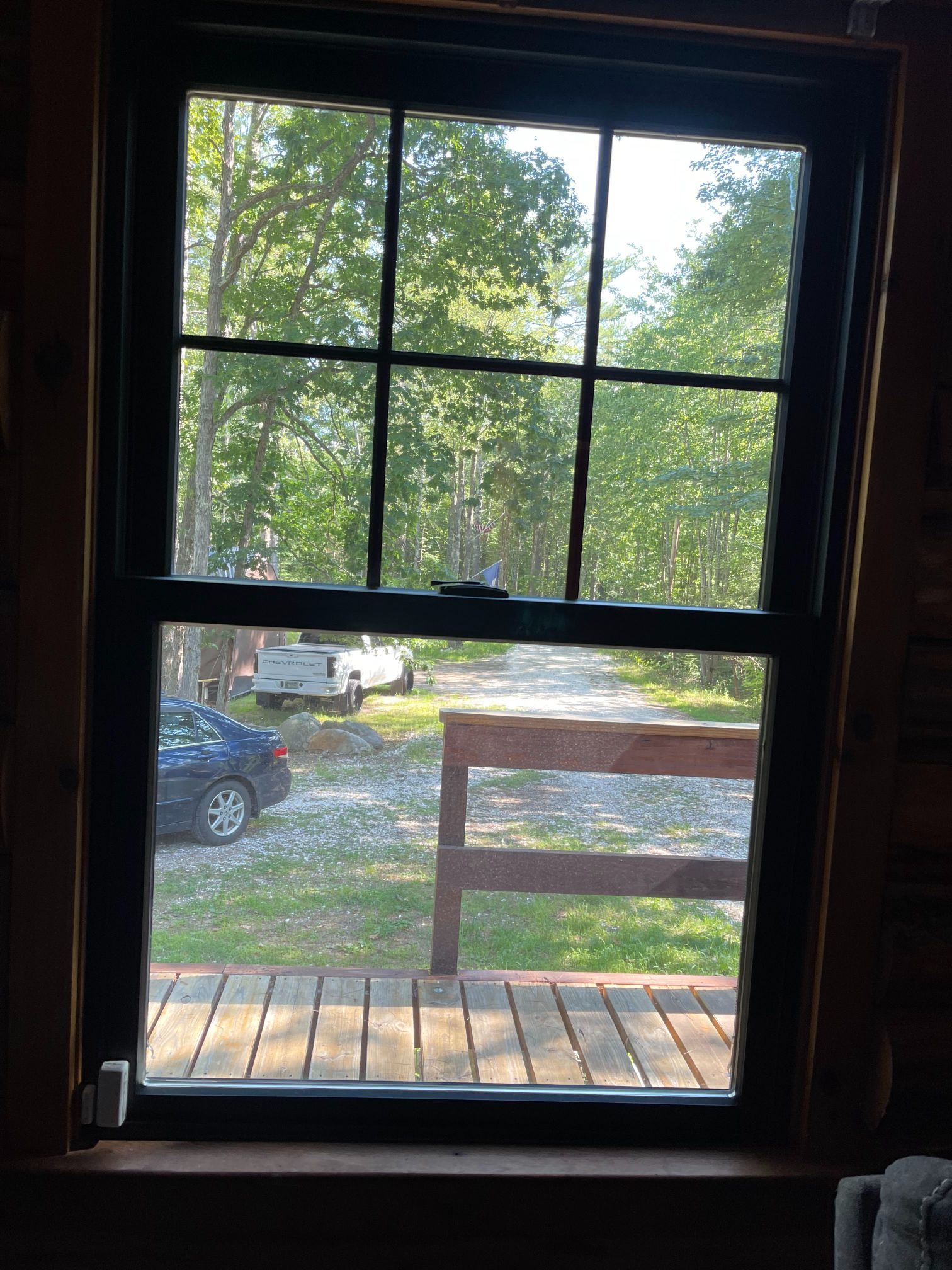 View through a black-framed window: a gravel driveway, cars, a wooden railing, and green trees.