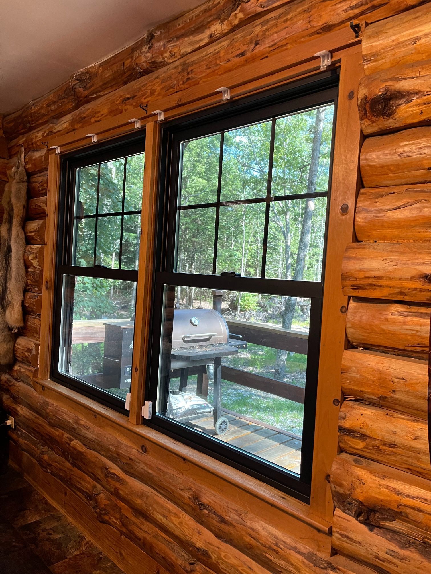 Two black-framed windows in a log cabin with a view of a grill, fence, and trees.
