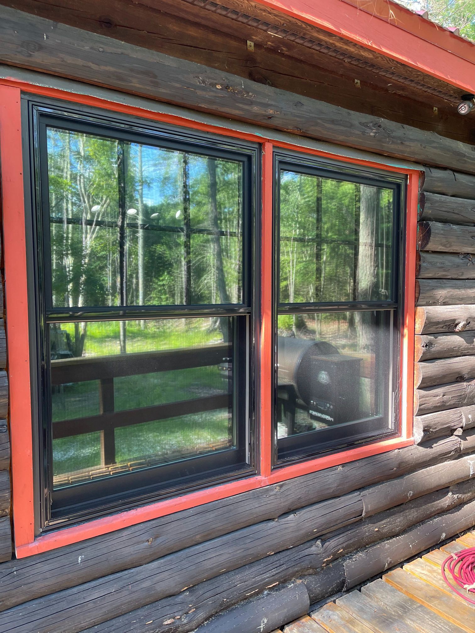 Two black-framed windows with red trim set in a log cabin wall, reflecting trees and a fence.