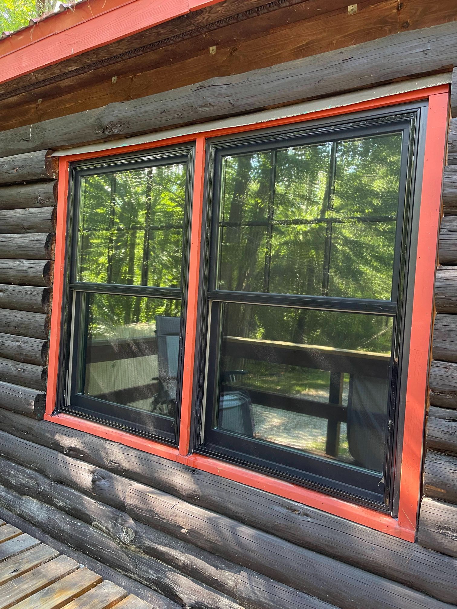 Two black-framed windows with red trim set into a log cabin wall, reflecting trees and a deck.