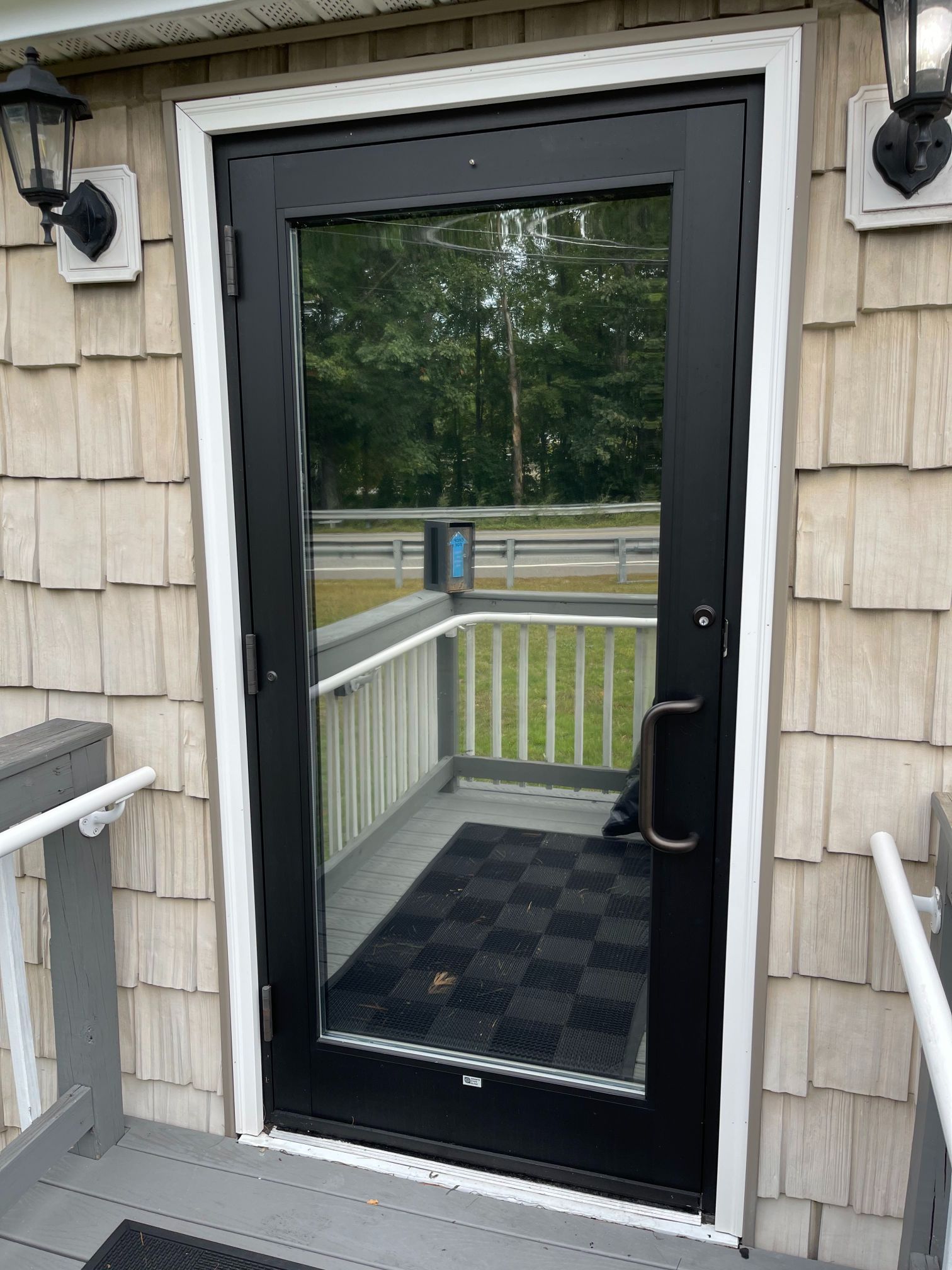 Black-framed glass door with a doormat, on a porch with gray railings. Siding is beige, with two black sconces.