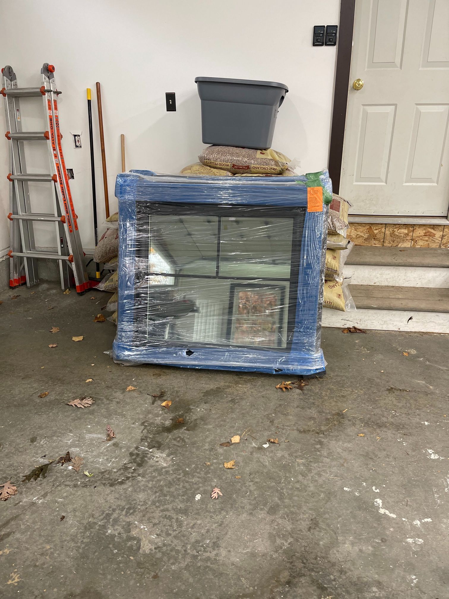 Window wrapped in blue plastic, on a garage floor. A ladder, trash bin, and door are in the background.