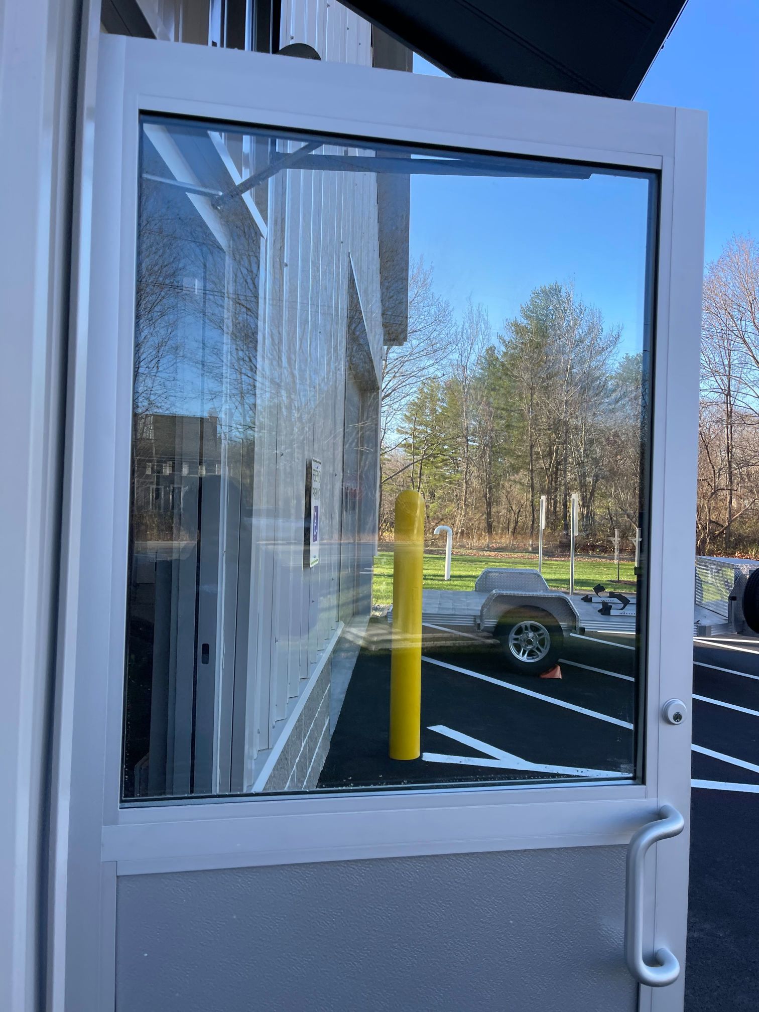 Open door with glass window reflecting a blue sky, trees, and a trailer in a parking lot.