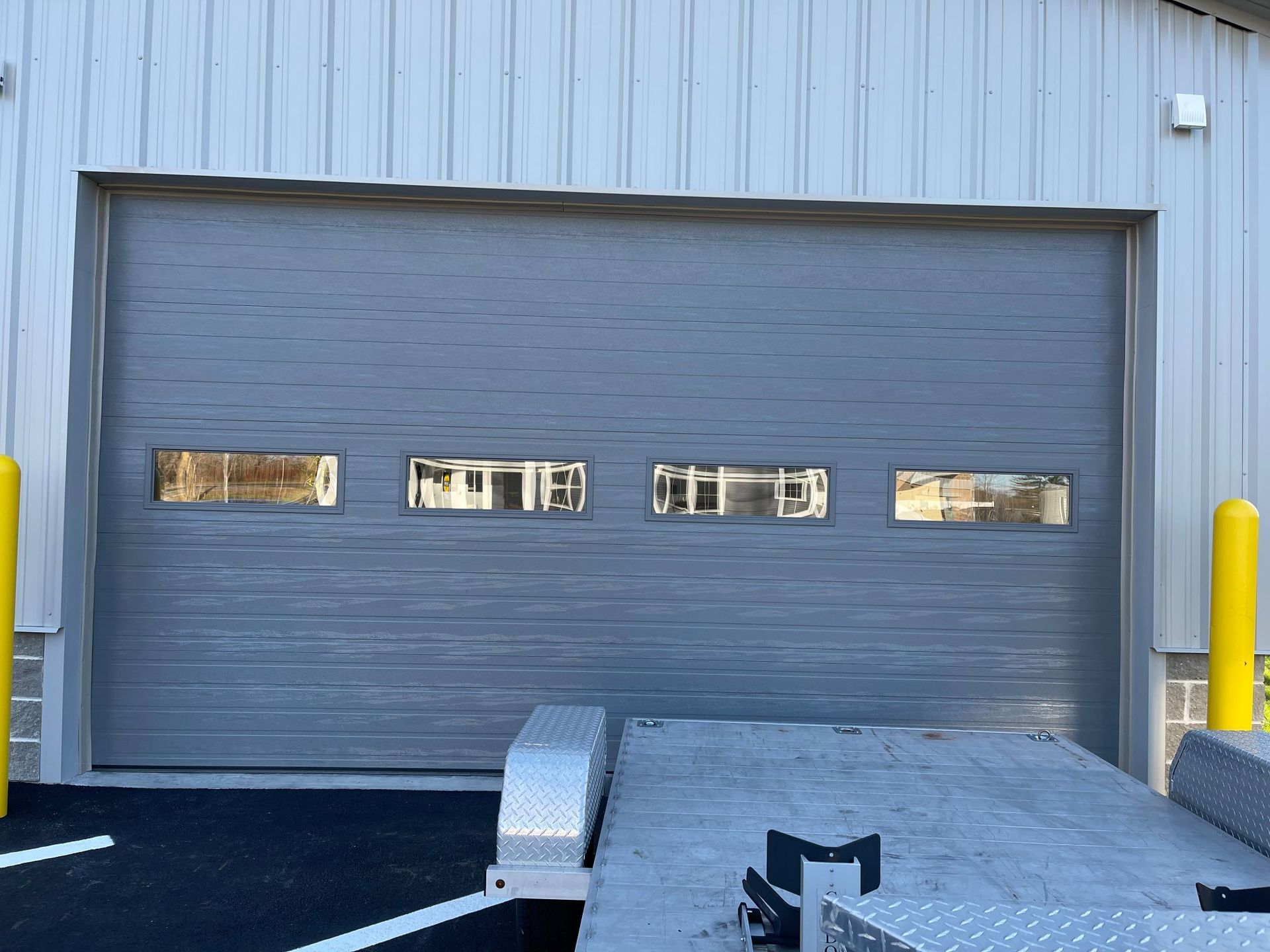 Gray commercial garage door with windows, facing a blacktop and a trailer. Yellow bollards flank the door.