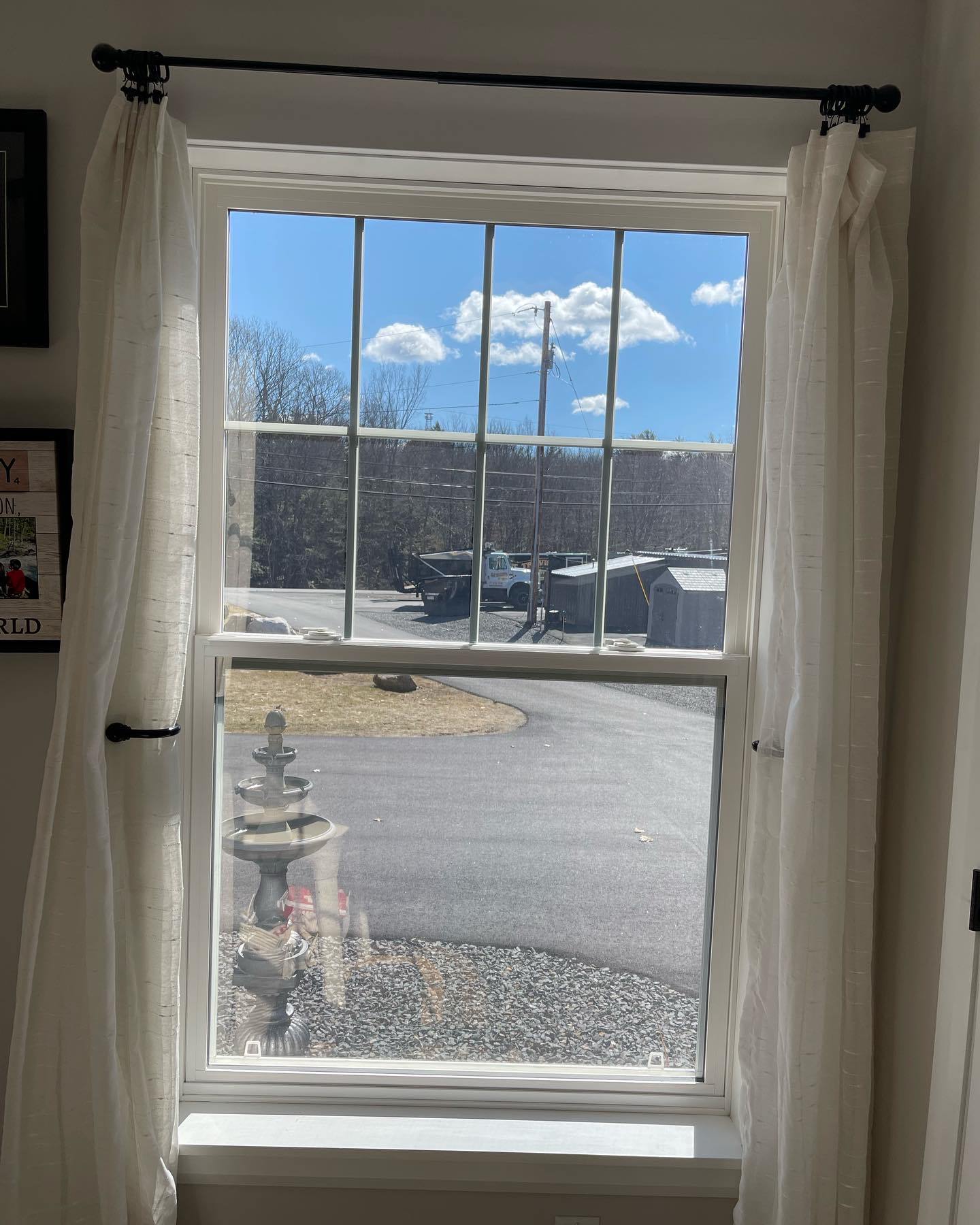 Window with sheer white curtains, revealing a sunny blue sky with clouds and a view of a street and buildings.