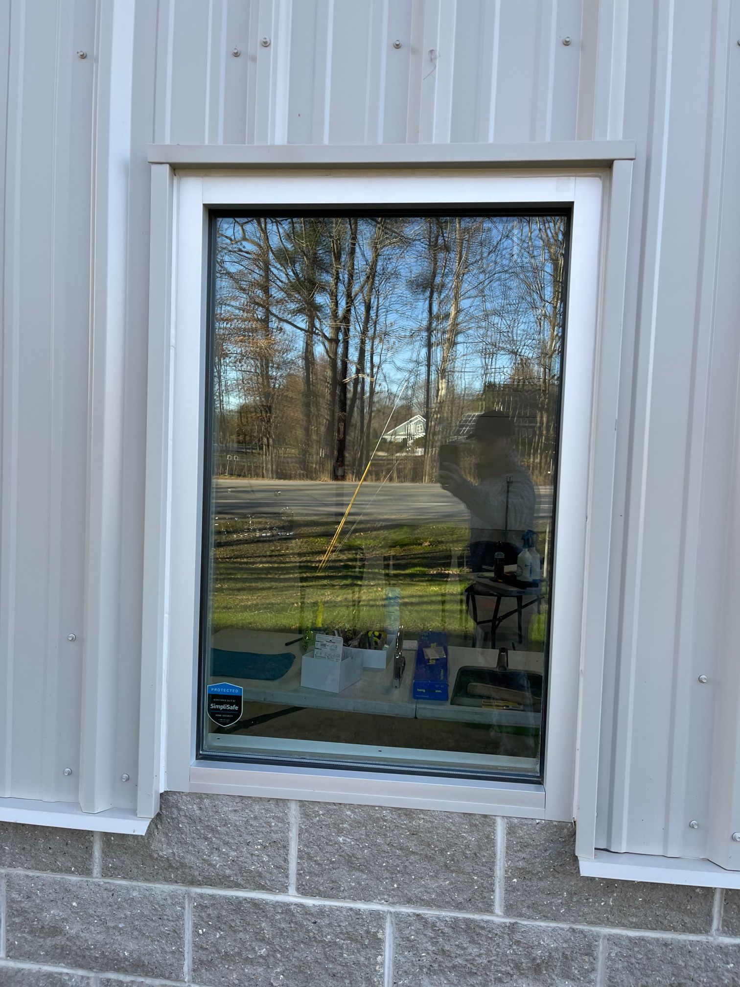 Window in a gray metal-sided building with reflection of trees and person. Window framed in white, set on cinder blocks.
