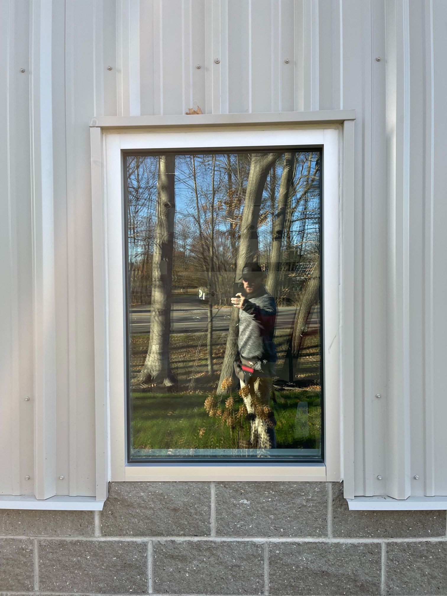 Window reflecting a person holding a phone, with trees and grass visible. The window is set in a tan metal building.