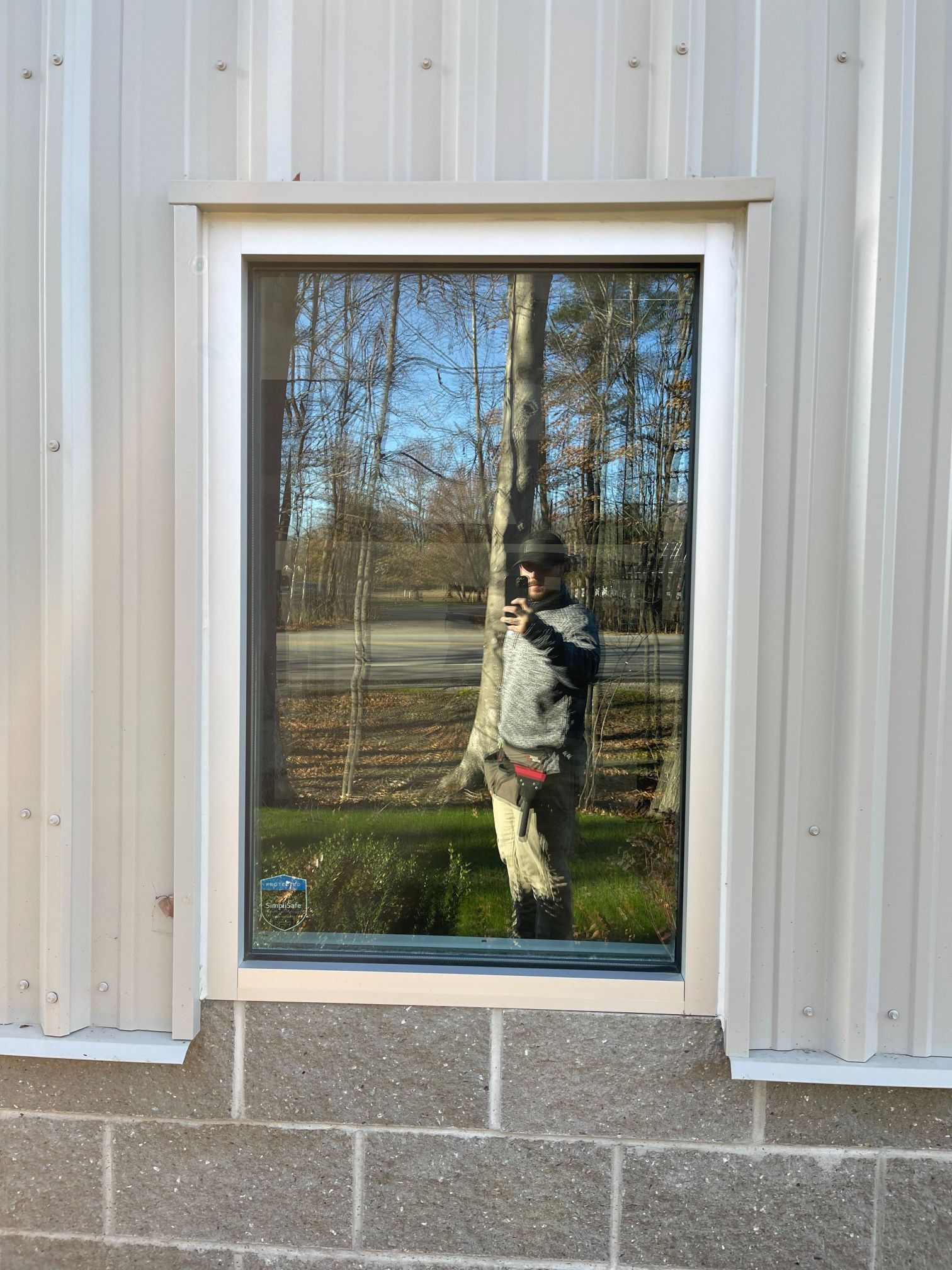 Window reflecting a person standing outdoors near trees, set in a building with beige metal siding over cinder blocks.