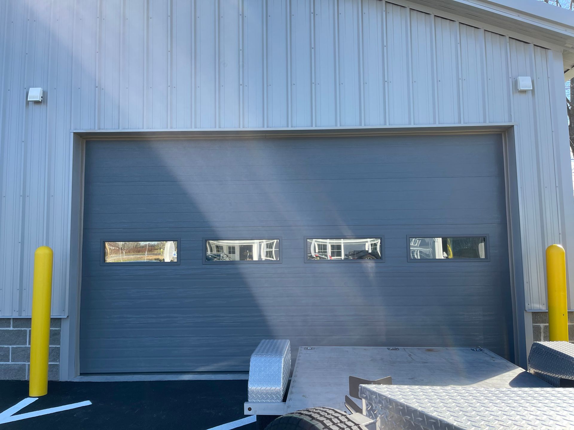 Gray garage door with three windows and yellow bollards. A trailer is in front.