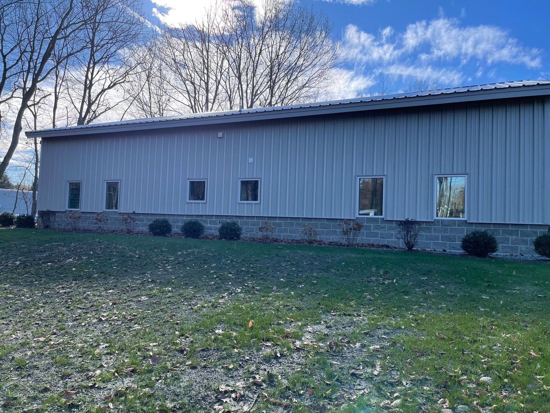 Long gray industrial building with several windows, set against a blue sky with trees.