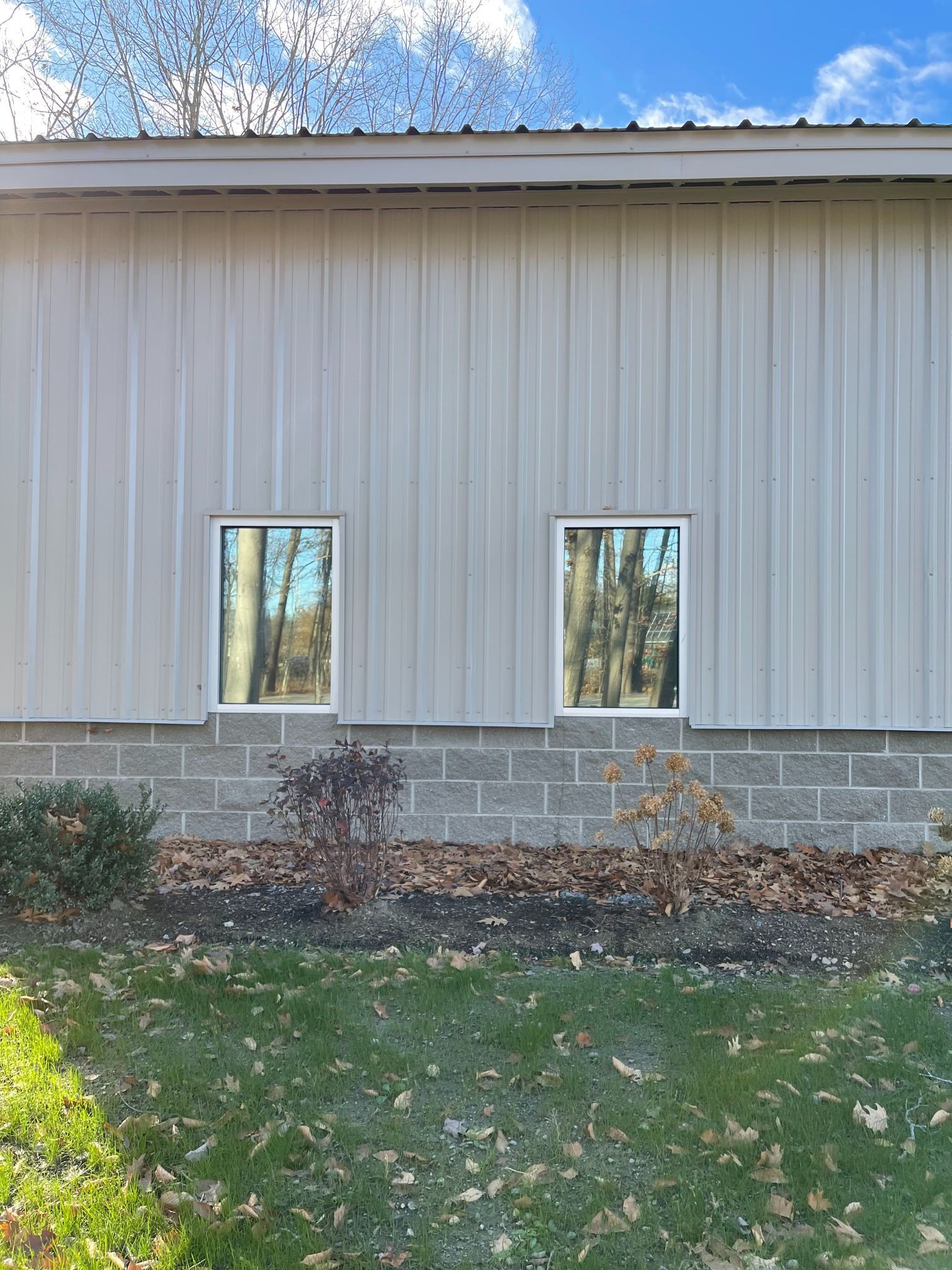 Exterior view of a building with two windows reflecting trees. Gray metal siding, brick foundation, and grass.