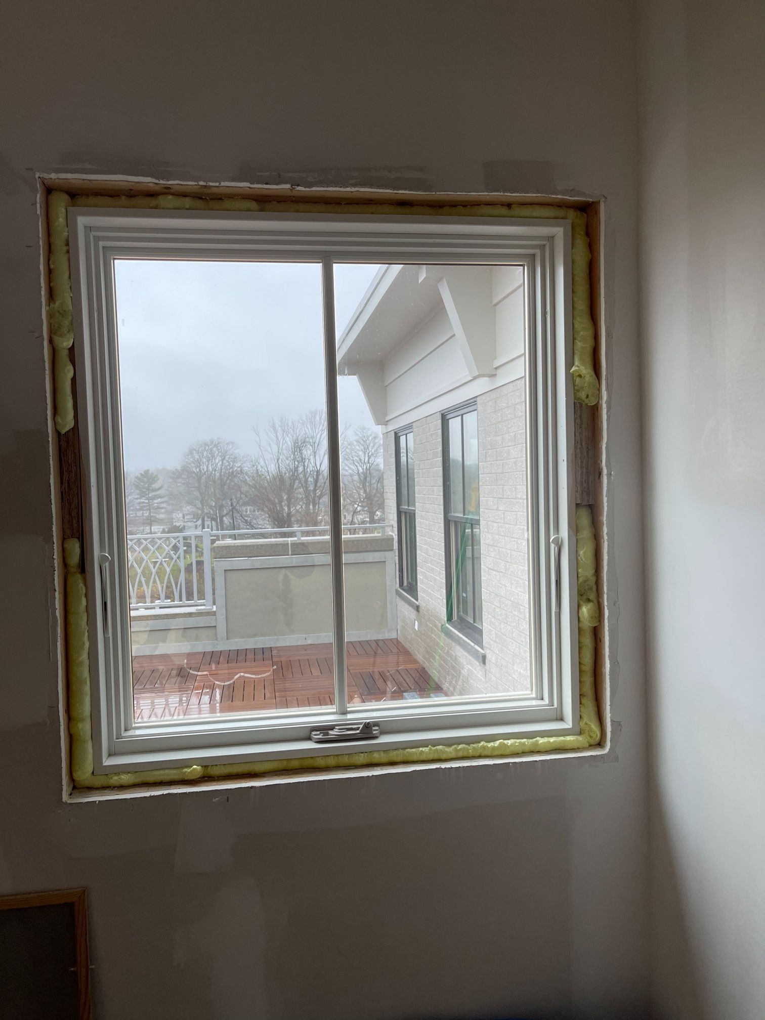 Square window, white frame, installed in a wall. Yellow foam insulation visible. Outside view of a house and snowy landscape.