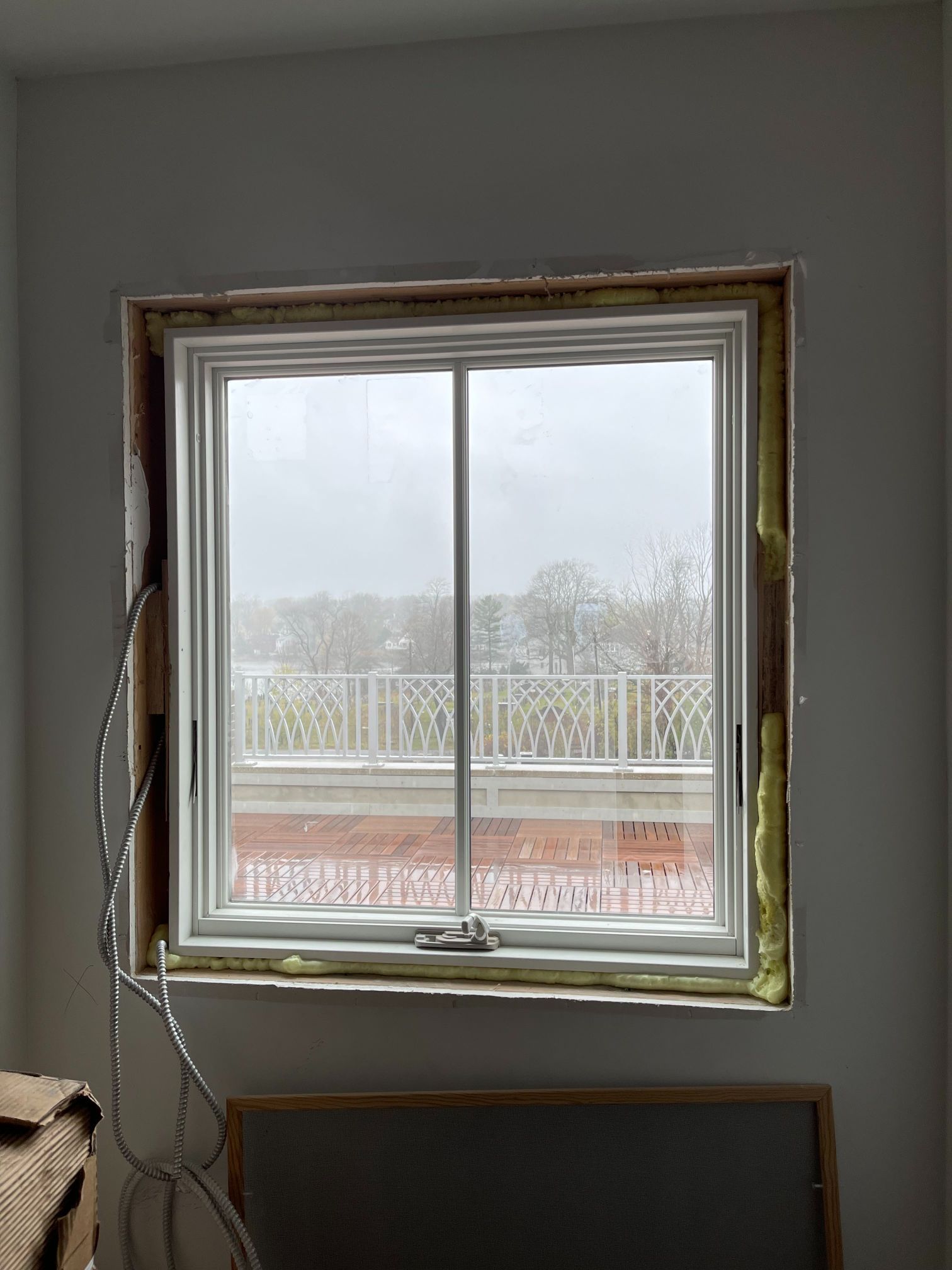 White window with outside view, installed in a wall with insulation visible. Wires hang nearby.