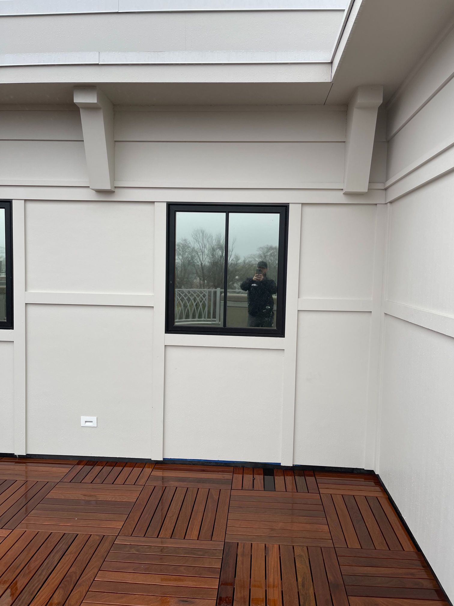 Wooden deck with white walls, a black-framed window, and a person reflected in the glass.