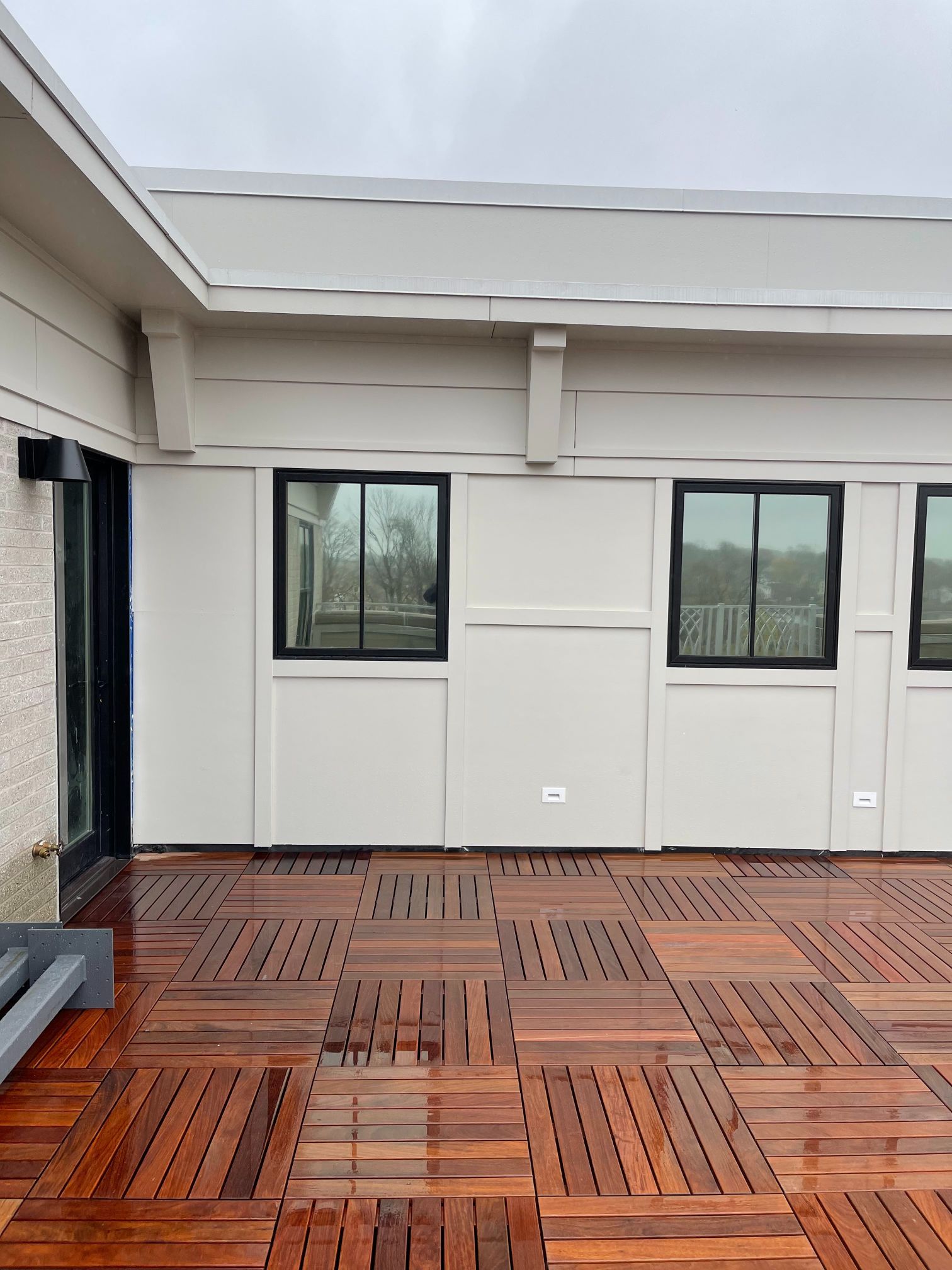 Wooden patio with a wall featuring black-framed windows under a light-colored roof overhang.