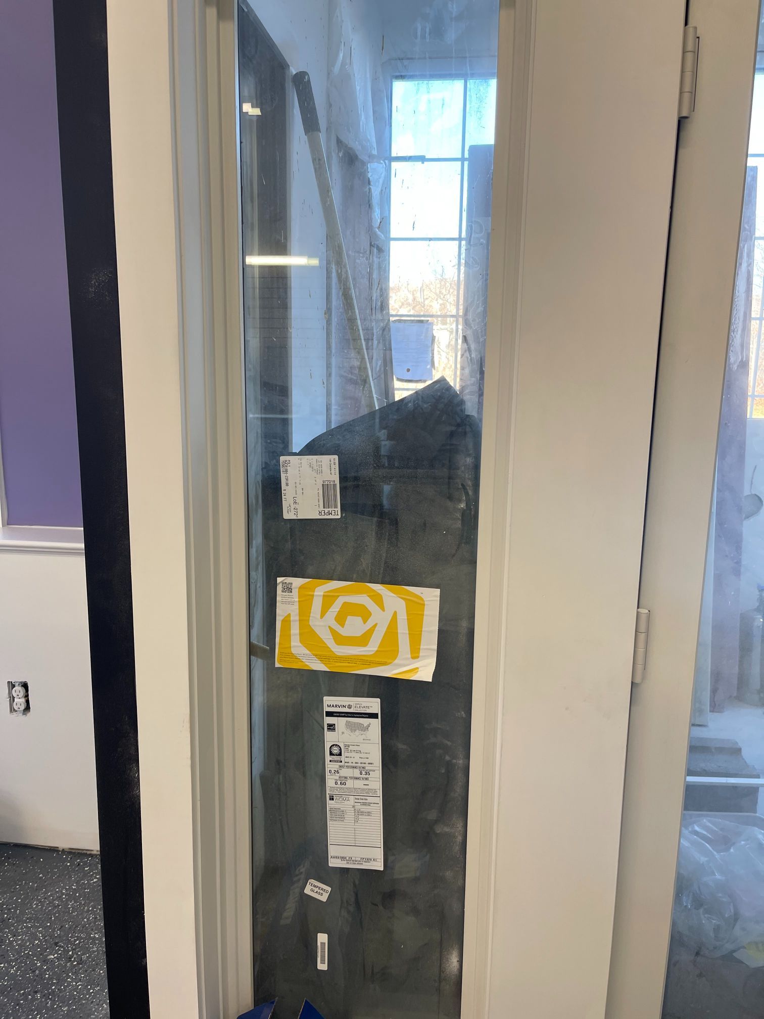 Window with black covering and yellow sticker, viewed from a room with purple wall.