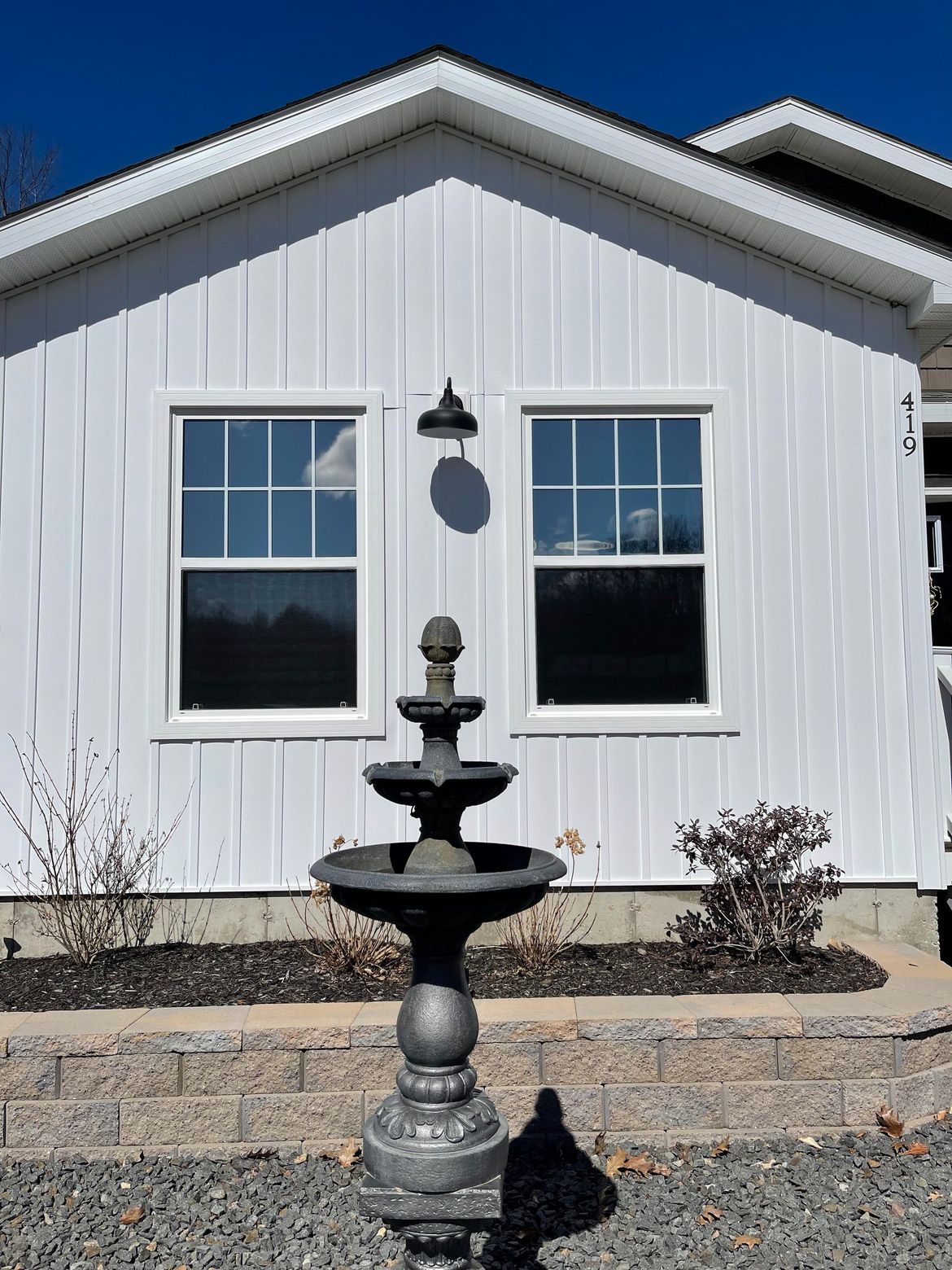 White house facade with two windows, a fountain, and gravel landscaping under a blue sky.