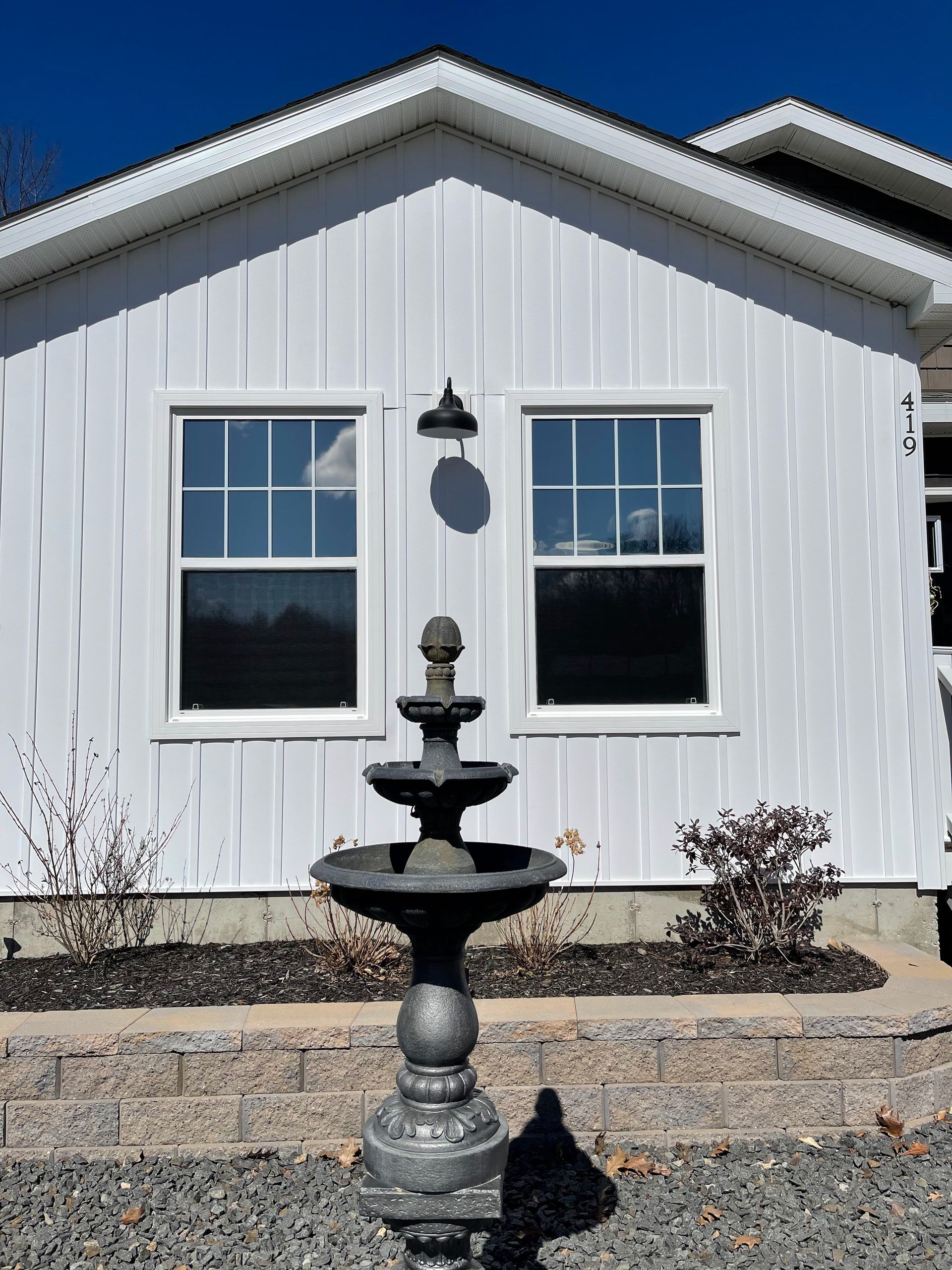 White house facade with two windows, a fountain, and gravel landscaping under a blue sky.