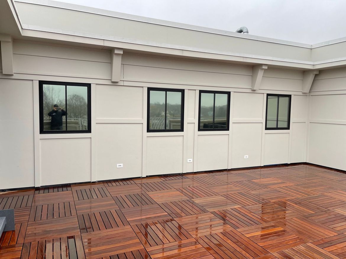 A partially covered outdoor patio with a series of black-framed windows in a beige wall, wooden floor tiles.