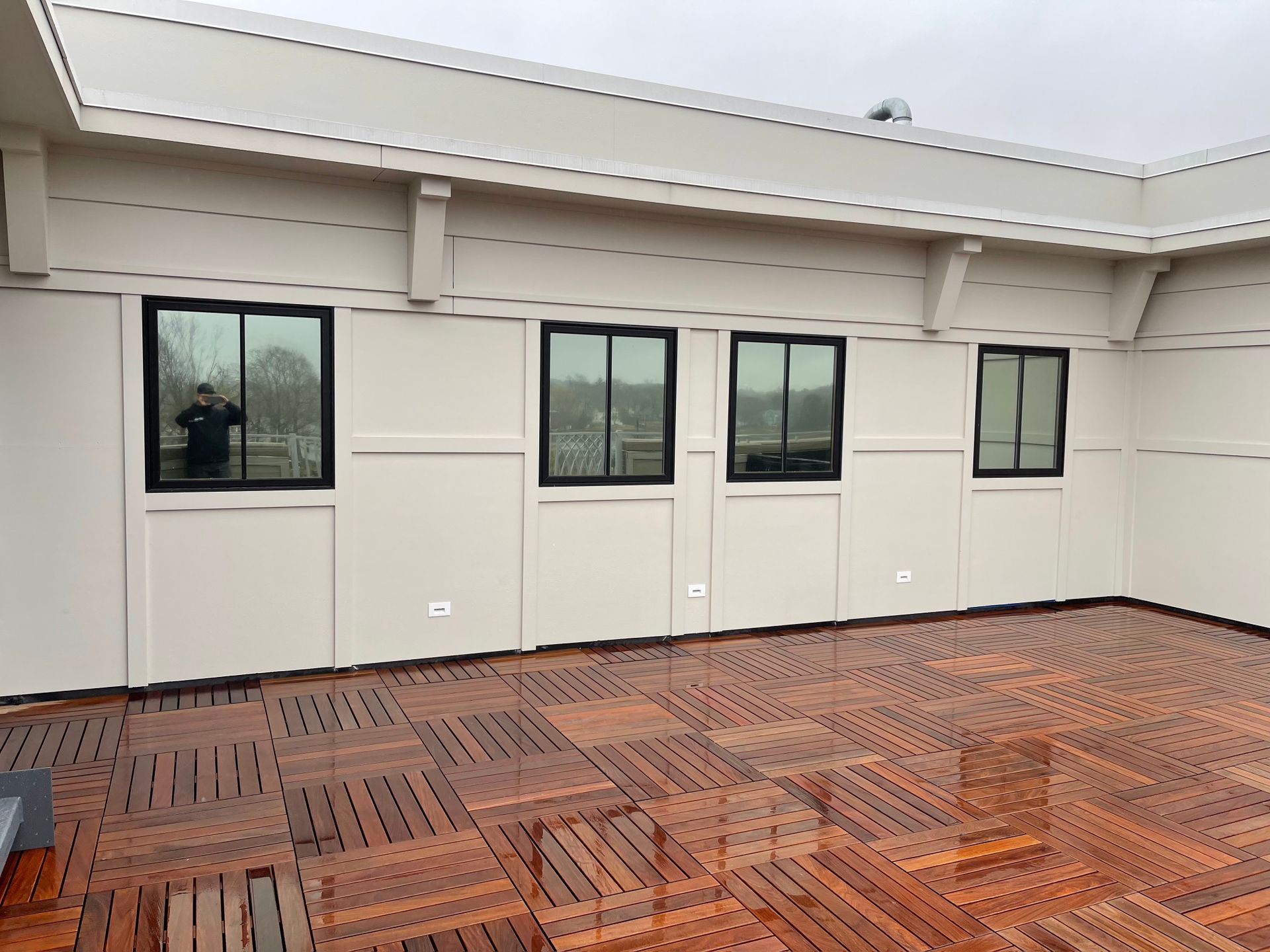 Exterior view: beige building with four black-framed windows, wet wooden floor.