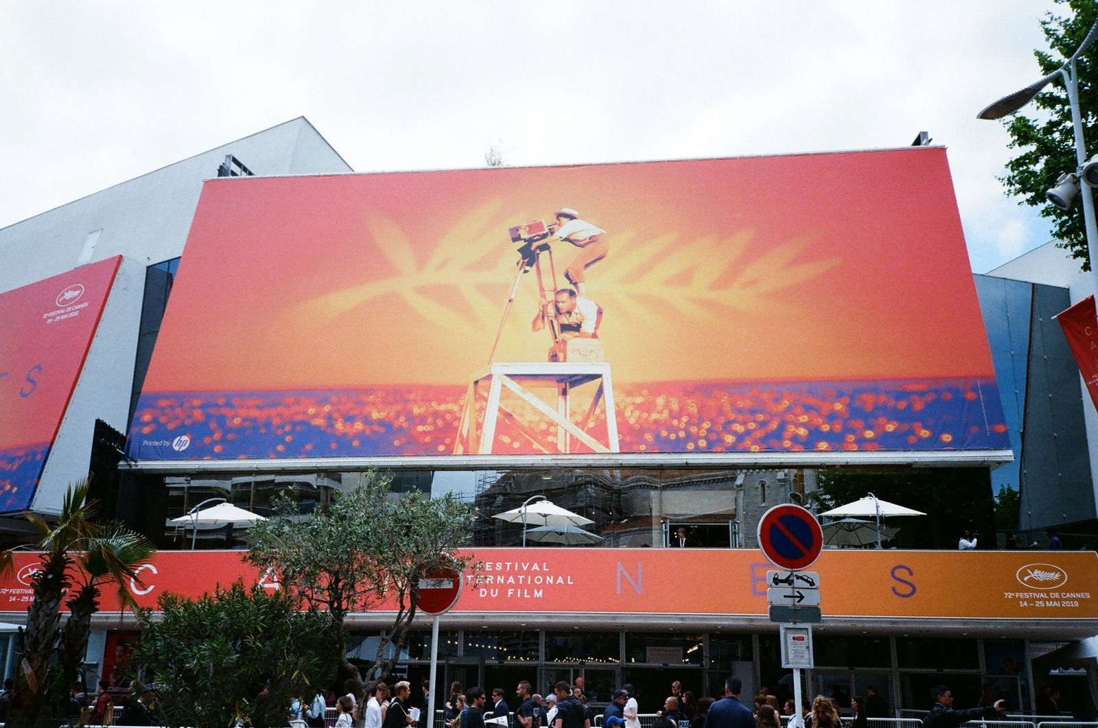 Cannes Film Festival building with large poster, sunset colors, figures with camera on a platform.