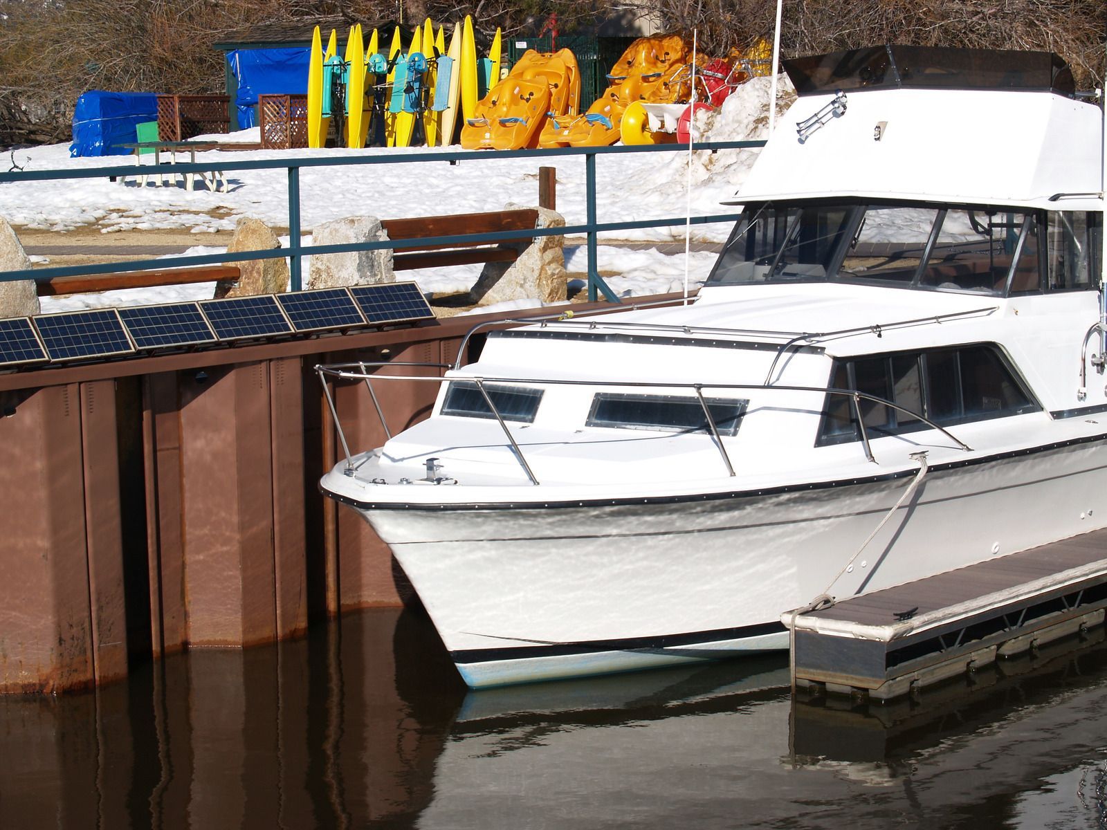 White boat docked at a pier, kayaks and gear visible behind it, winter setting.