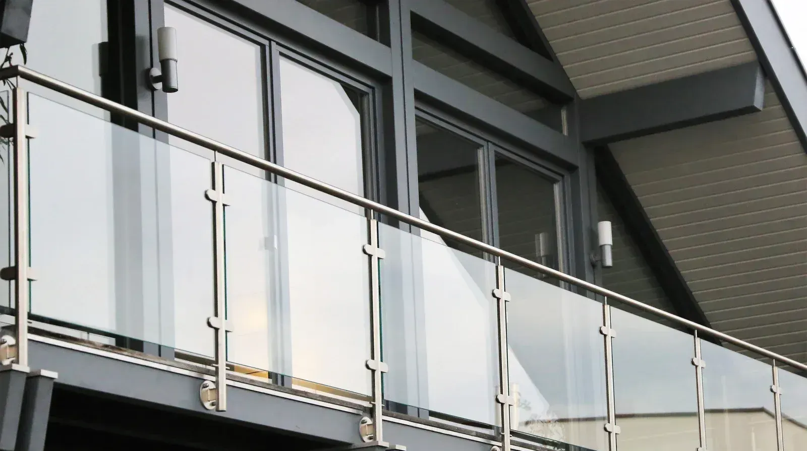 Balcony with glass panels and stainless steel railing. Building with dark window frames and angled roof.
