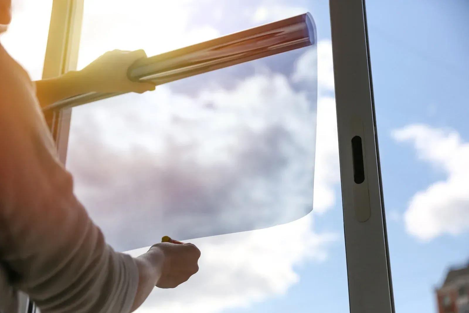 Person applying window film, holding the sheet against the glass, sunny sky visible.