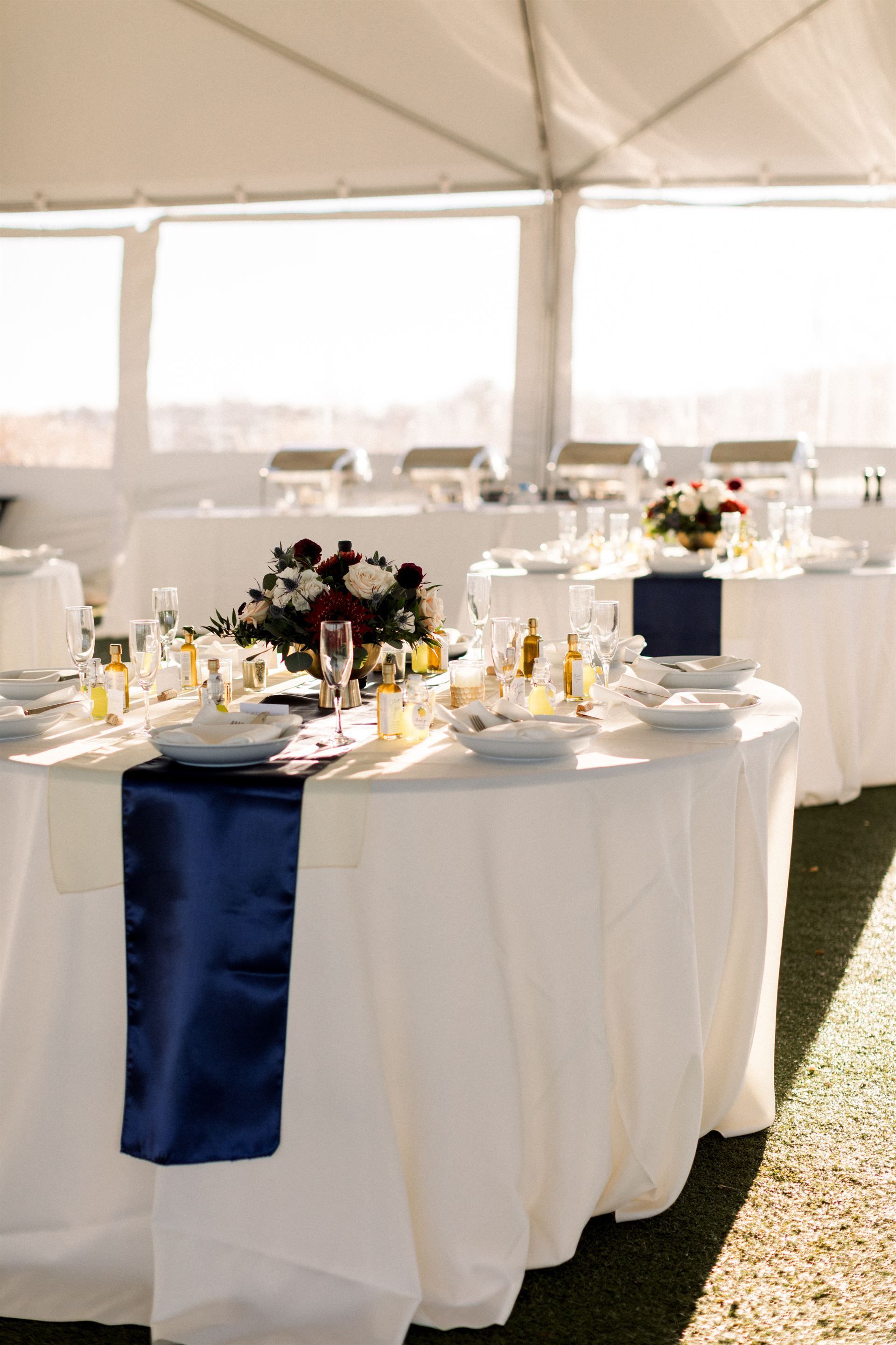 Wedding reception tables in tent, white tablecloths, navy runner, centerpieces, catering dishes in background.