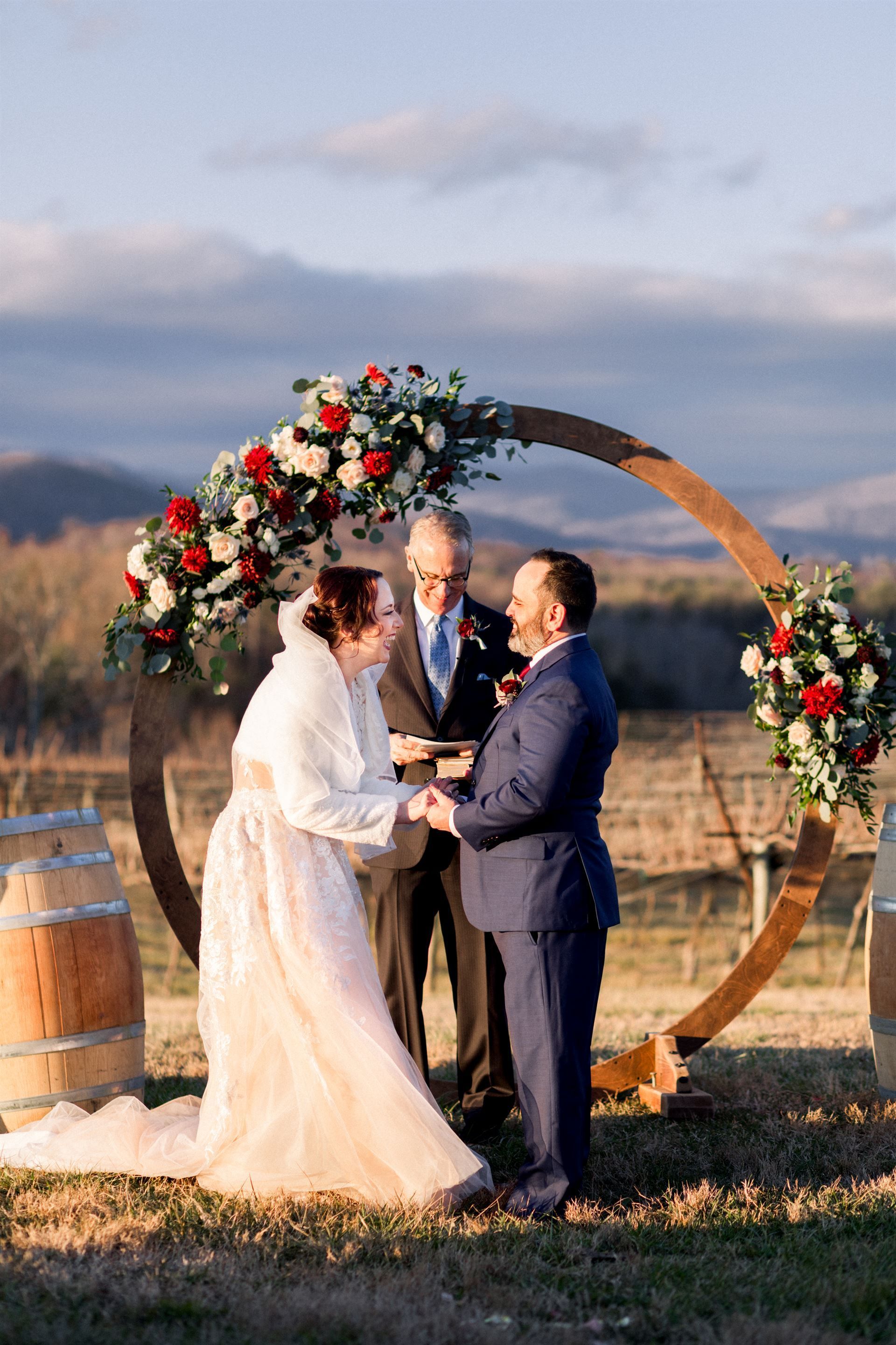 Couple exchanging vows at outdoor wedding, wooden arch with red and white flowers, officiant, vineyard backdrop.