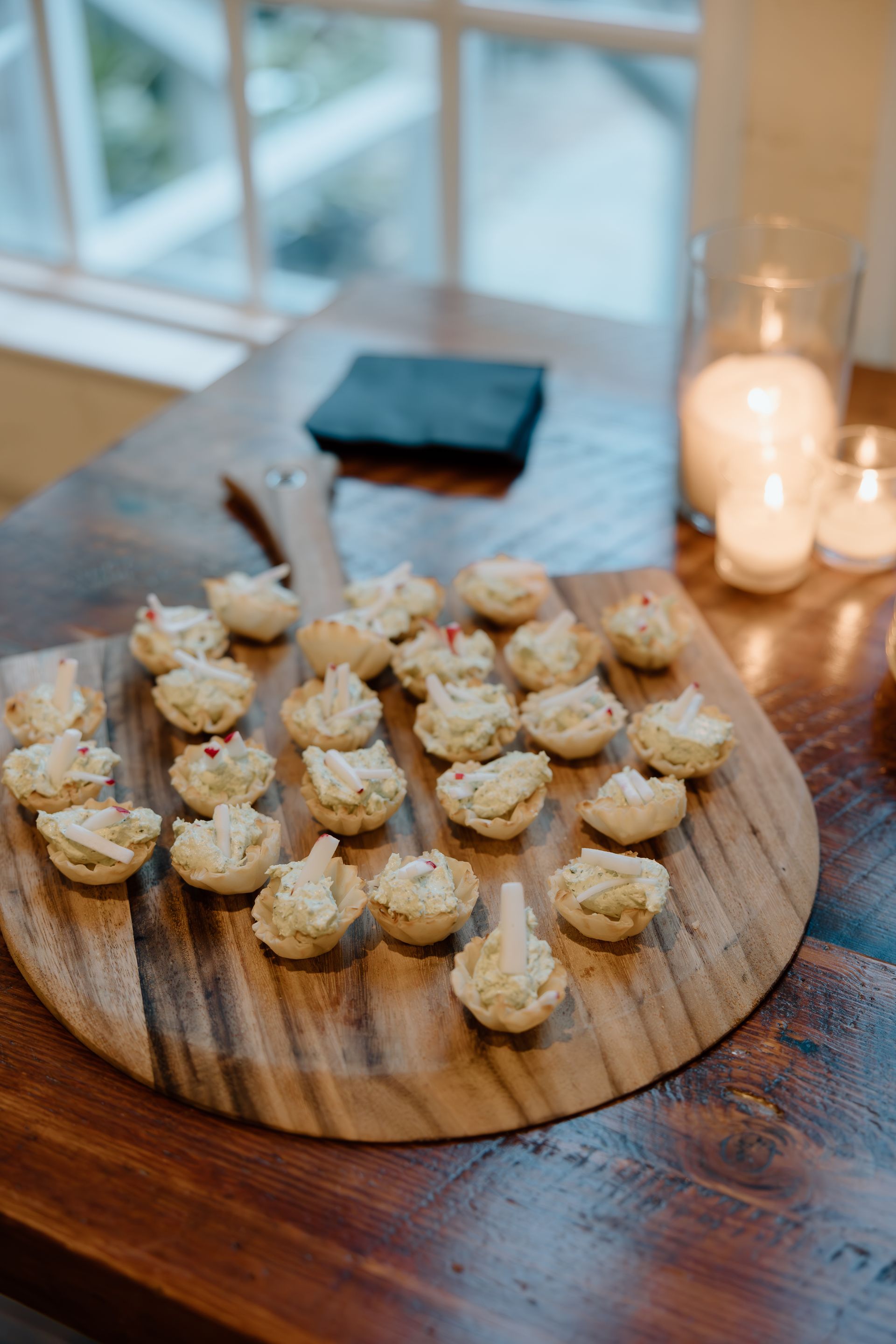Appetizers on a wooden paddle, near a window and candles.
