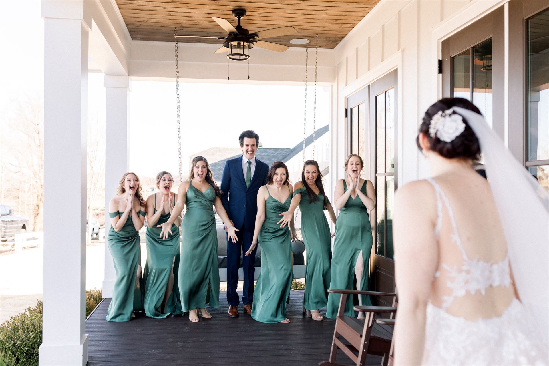 Bride on porch facing bridesmaids and groom, all reacting with surprise. They wear green dresses.