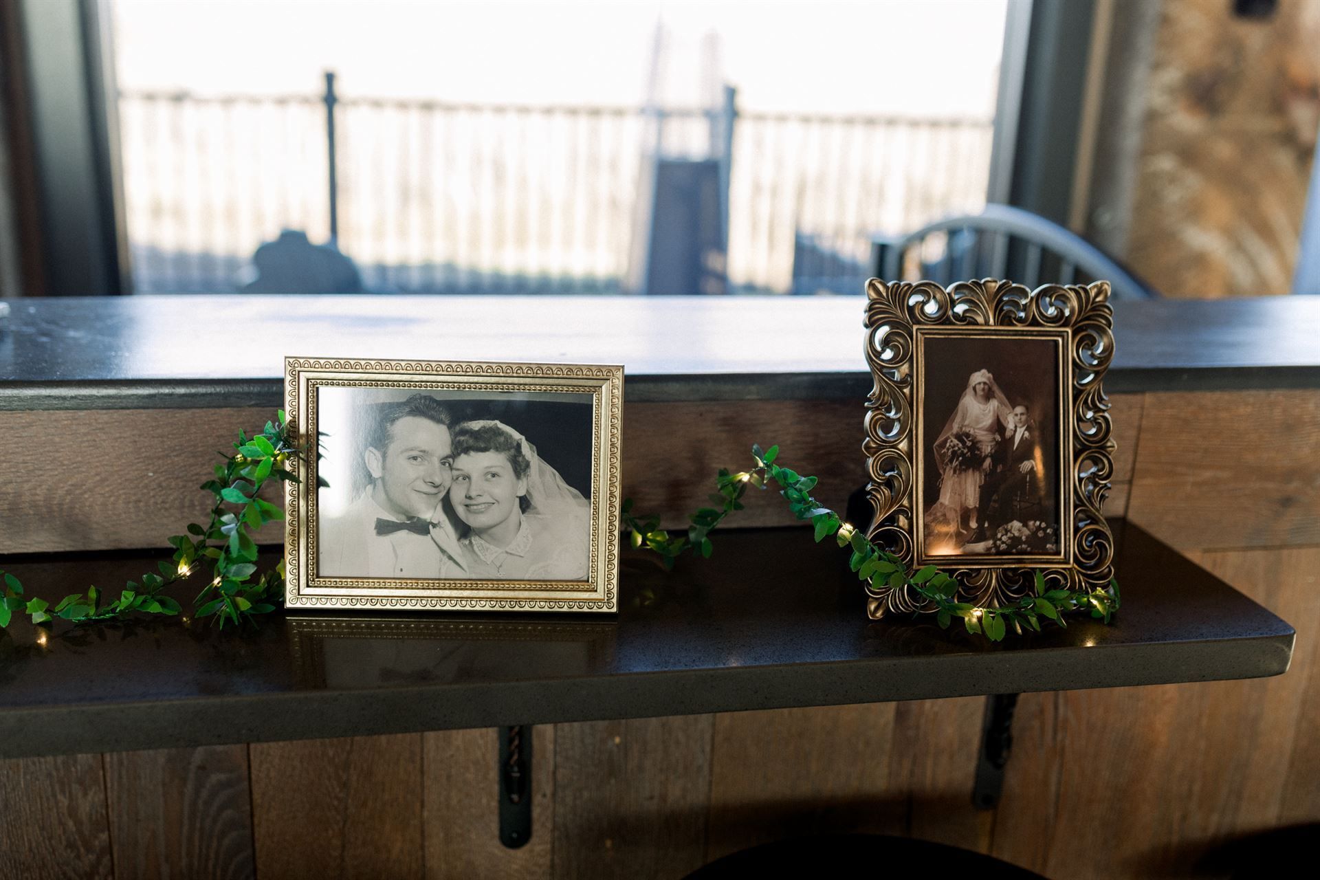Two framed vintage photos sit on a wooden bar, decorated with greenery and lights.