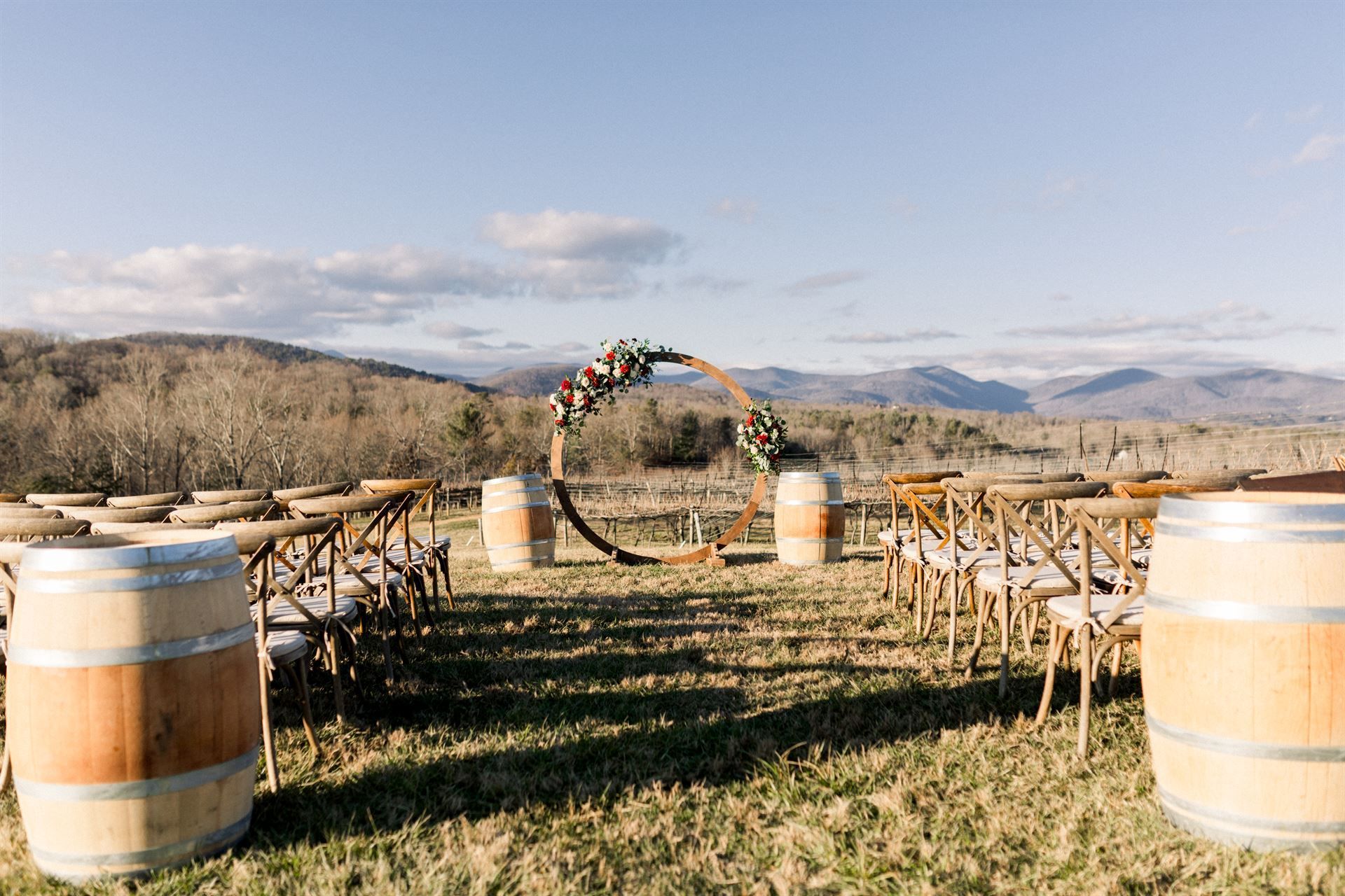 Wedding ceremony setup in field with arch, barrels, chairs, and mountain backdrop.