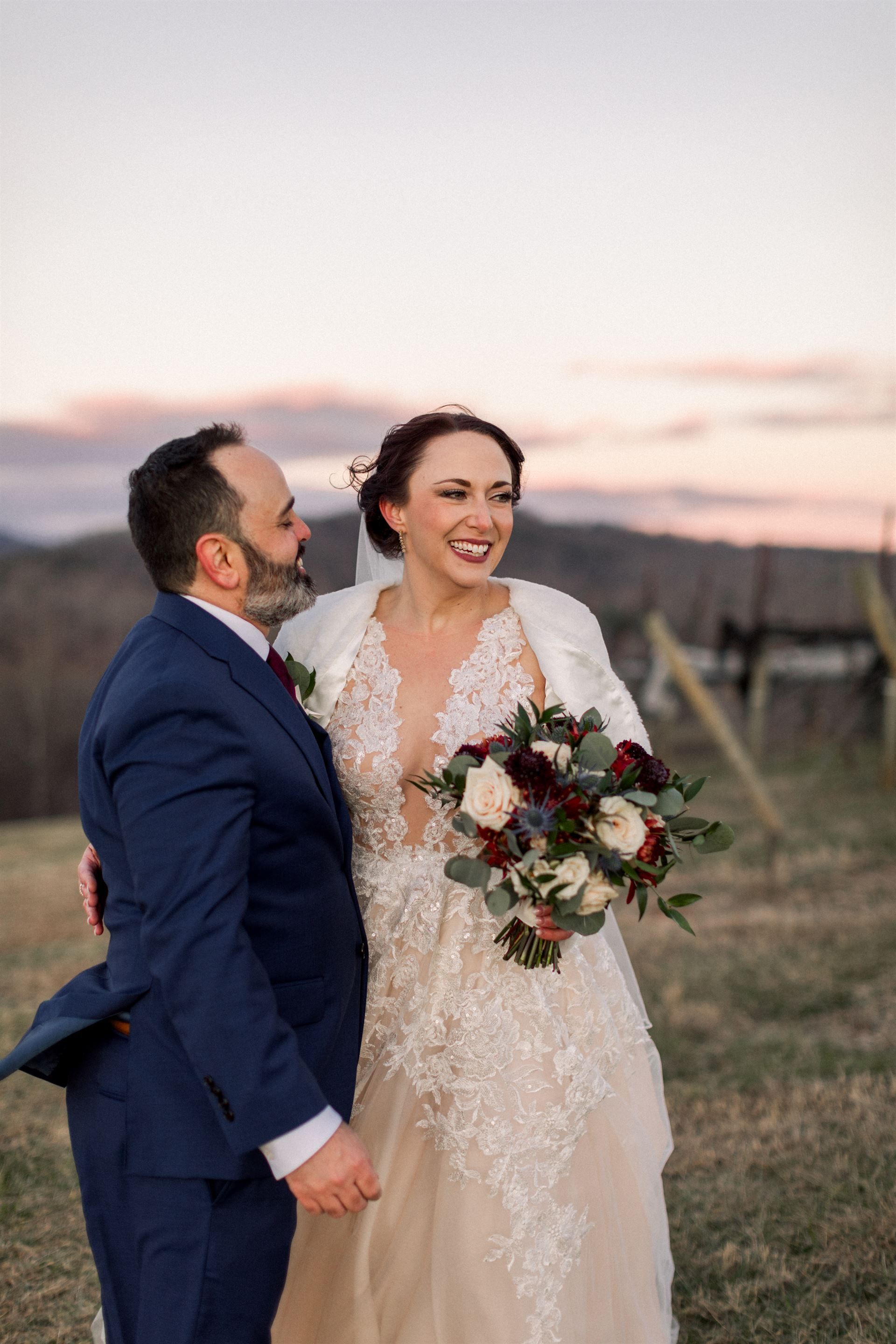 Bride and groom embrace outdoors at sunset; she smiles, holding bouquet, he looks at her, both in formal attire.