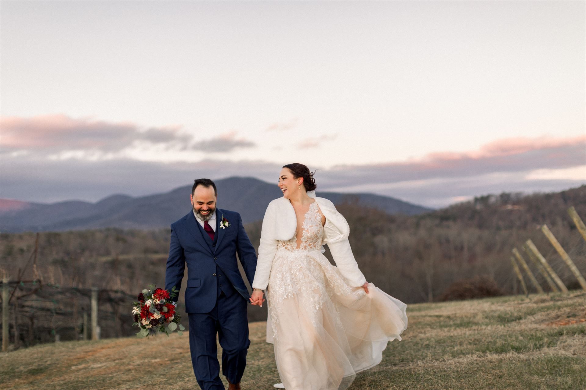 Bride and groom walk hand-in-hand, smiling, across a grassy hill. Mountain backdrop, dusk light.