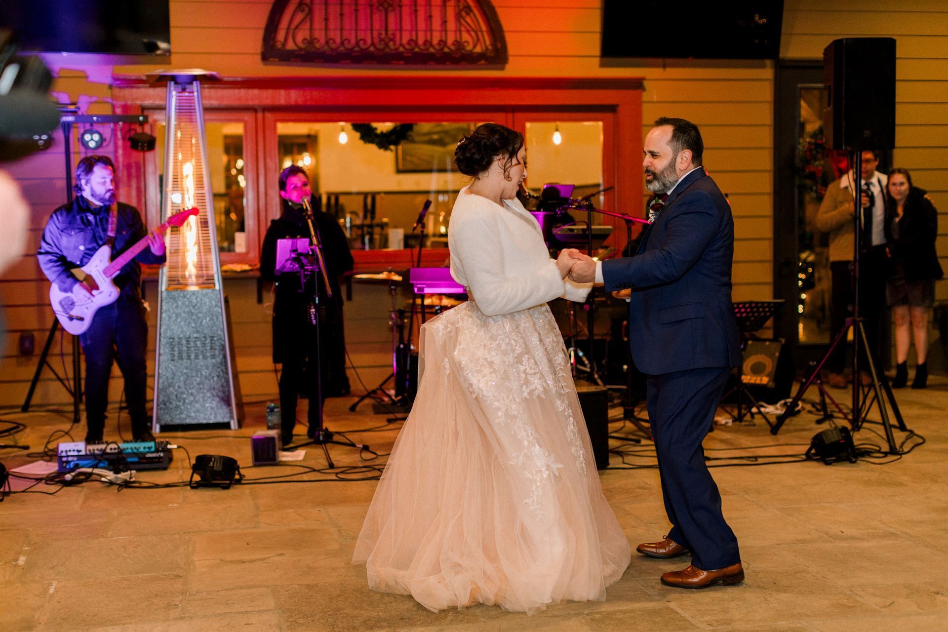 Bride and groom dance at reception with live band on stage. Warm lighting.