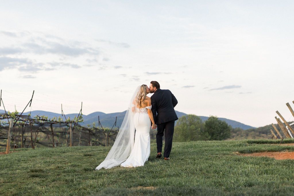 Bride and groom walk, groom kisses bride's cheek, in a vineyard with mountains in the background.