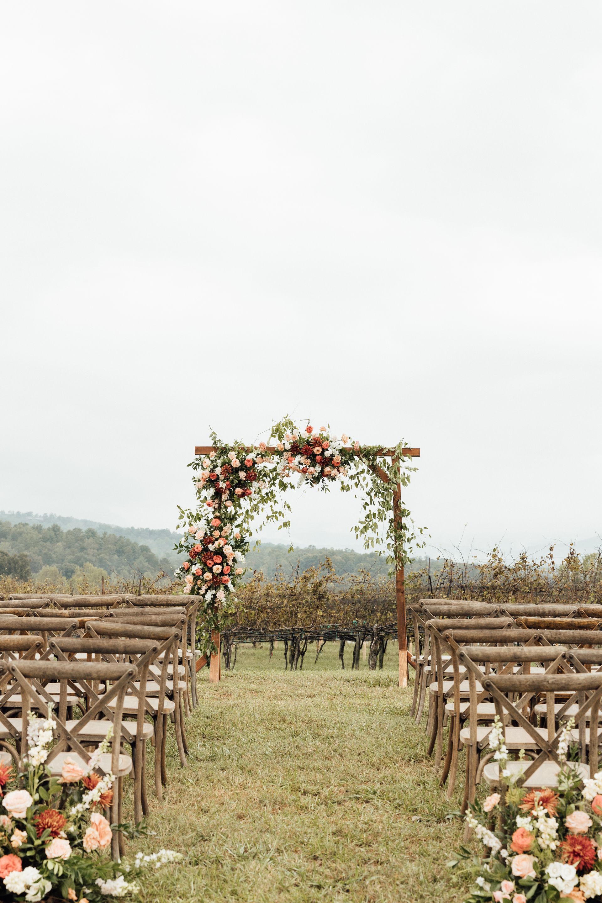 Wedding ceremony setup outdoors: wooden arch with floral decorations, rows of chairs face the altar, vineyard backdrop.