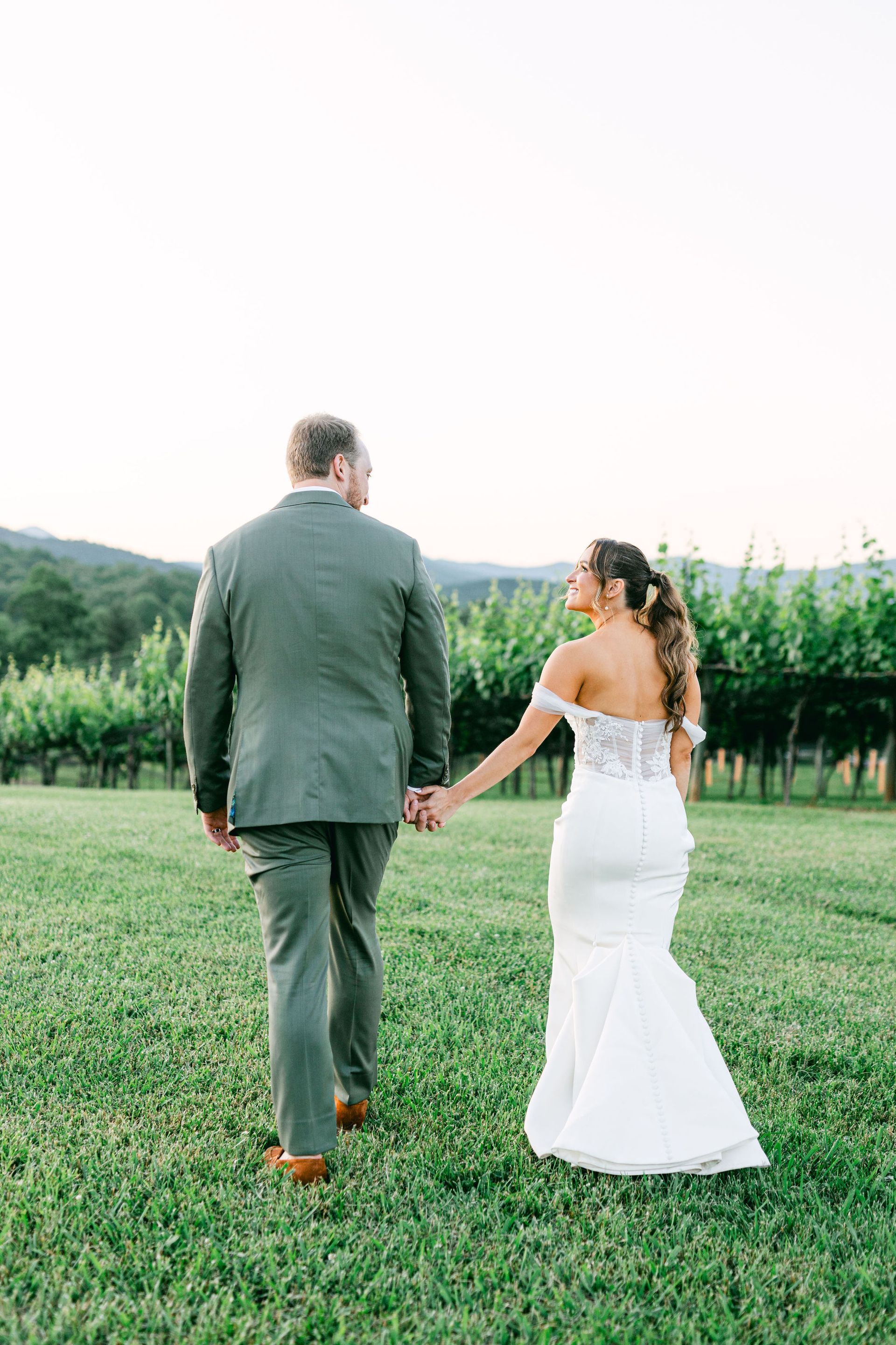 Bride and groom walking hand-in-hand through a vineyard. The bride wears a white gown, the groom a green suit.